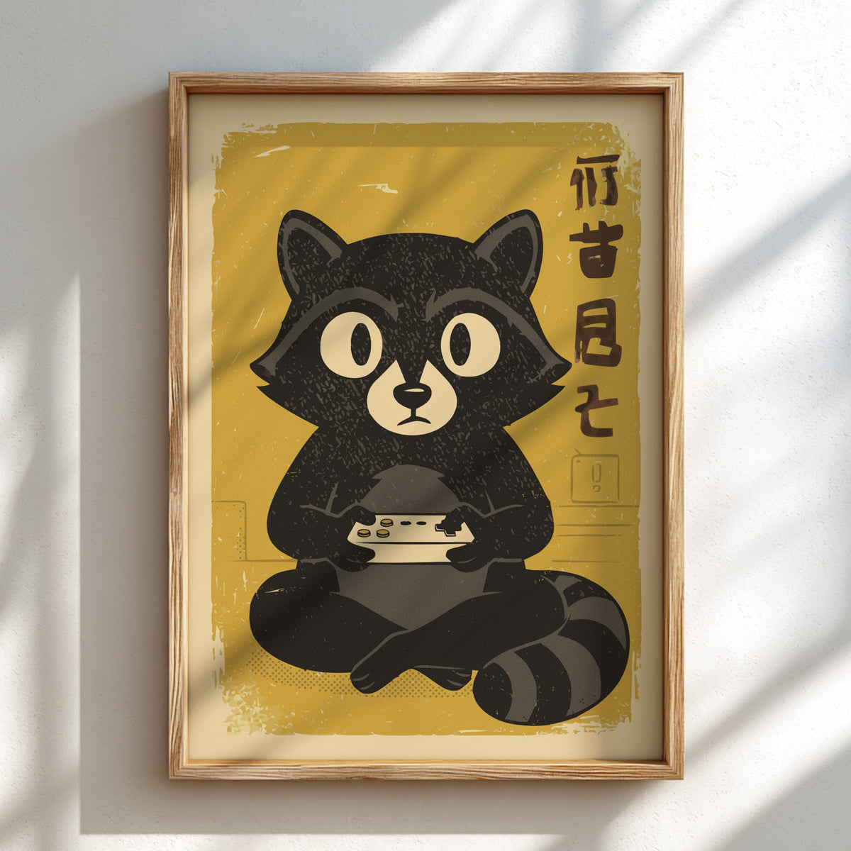 A framed art print featuring a stylized black raccoon holding a game controller on a yellow background, with vertical black characters on the right. The print is housed in a light wood frame and displayed on a white wall with soft diagonal shadows.