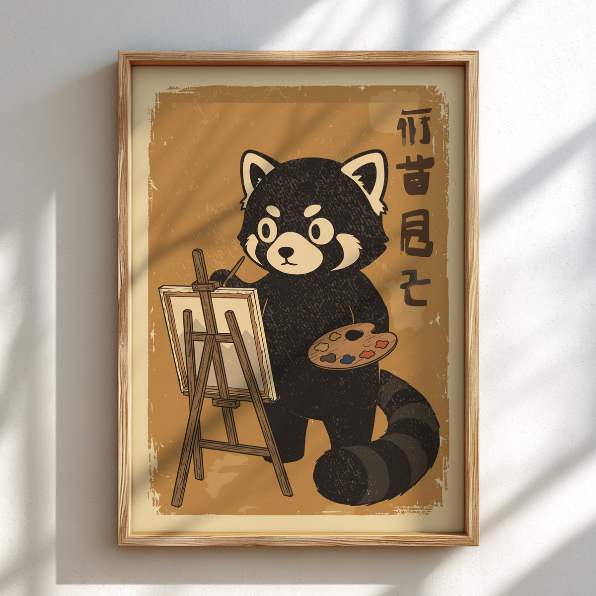 A framed art print featuring a cartoon red panda painting at an easel, displayed on a light wall with soft window shadows. The print has a warm tan background with black and cream details, enclosed in a light wood frame.