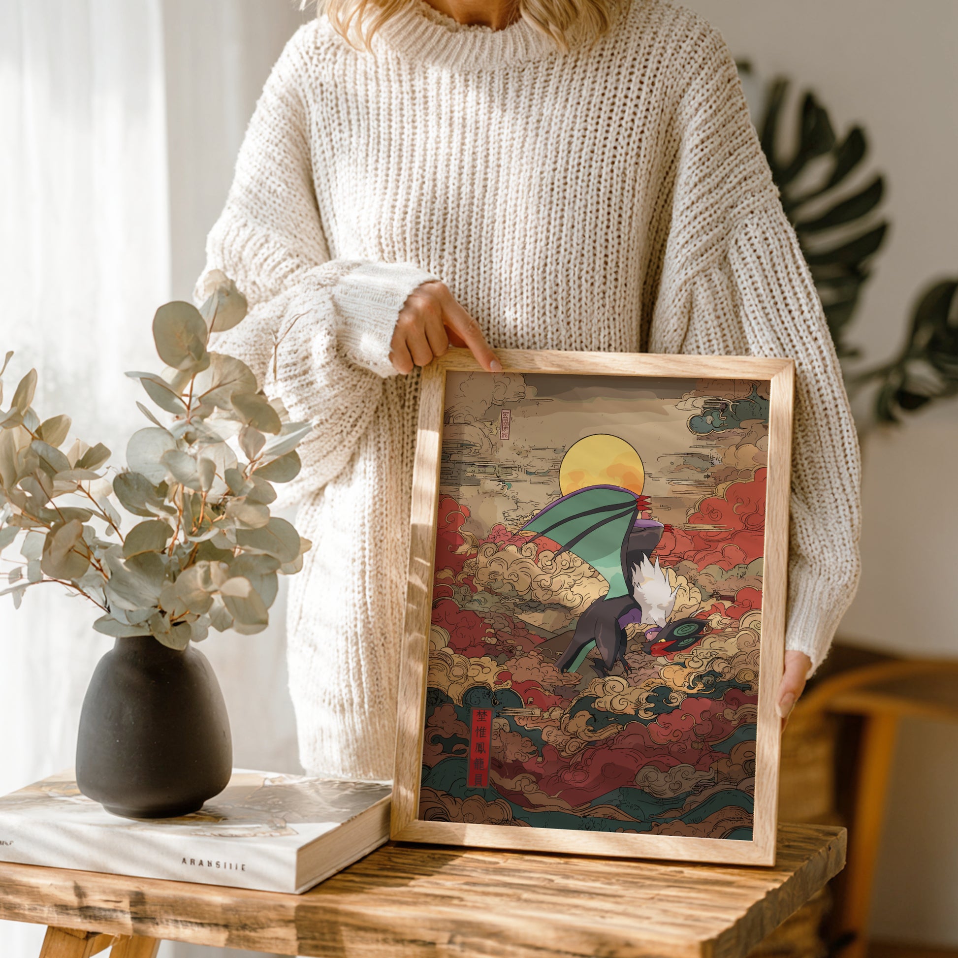 Person holding a framed artwork with a colorful design of Noivern Pokemon, standing next to a vase and book on a wooden table.