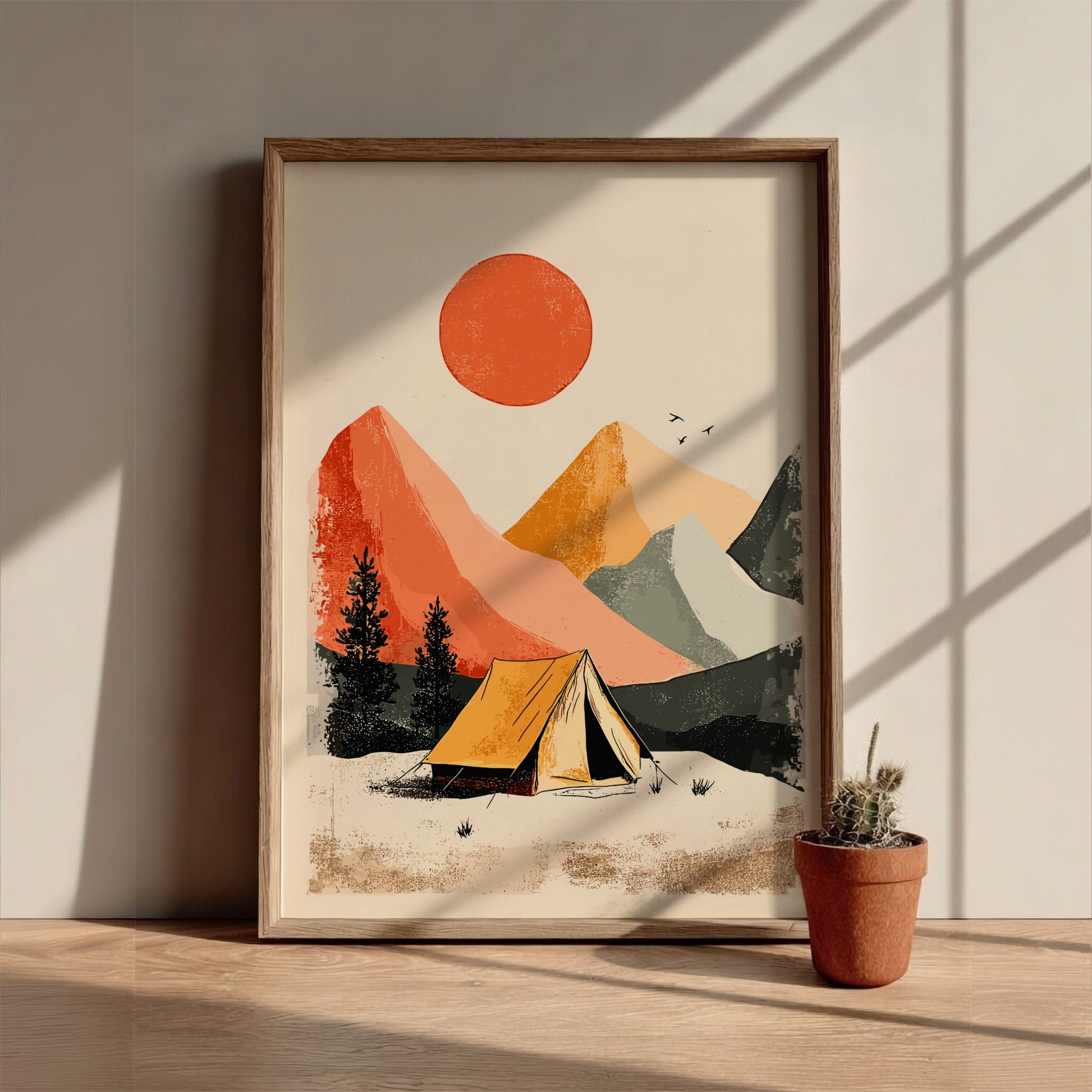 Framed artwork of a mountain landscape with a tent and sun, on a wooden surface with a plant.
