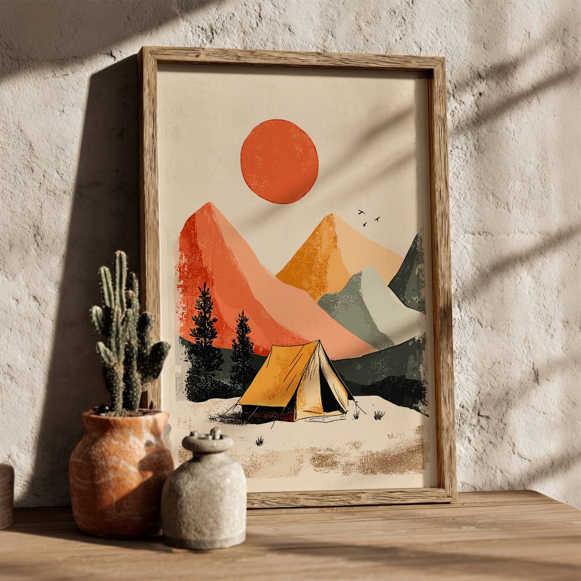 Framed artwork of a mountain landscape with a tent and sun, on a wooden surface with a plant.