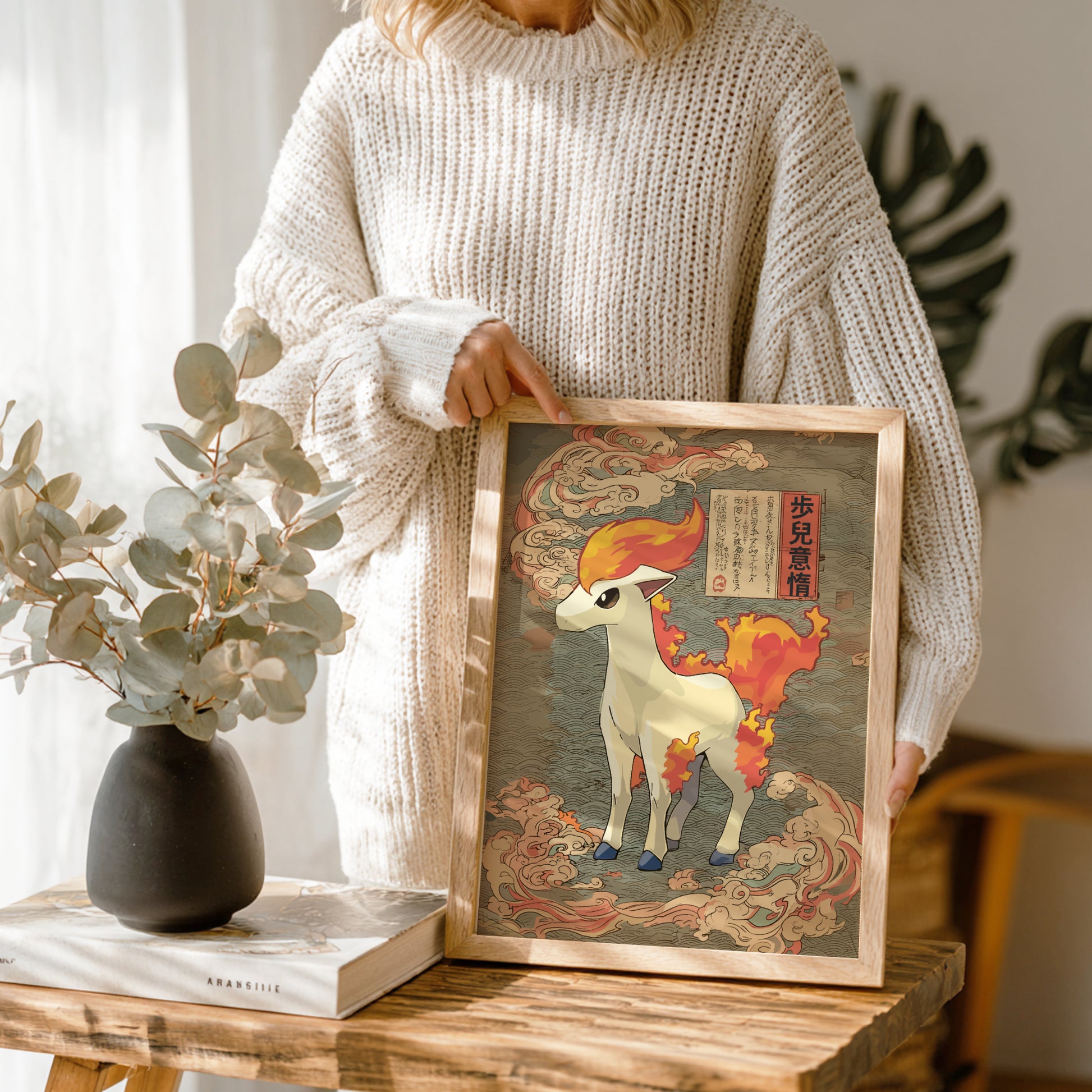 Person holding a framed artwork of Ponyta Pokemon on a wooden table with a vase and book in the background.