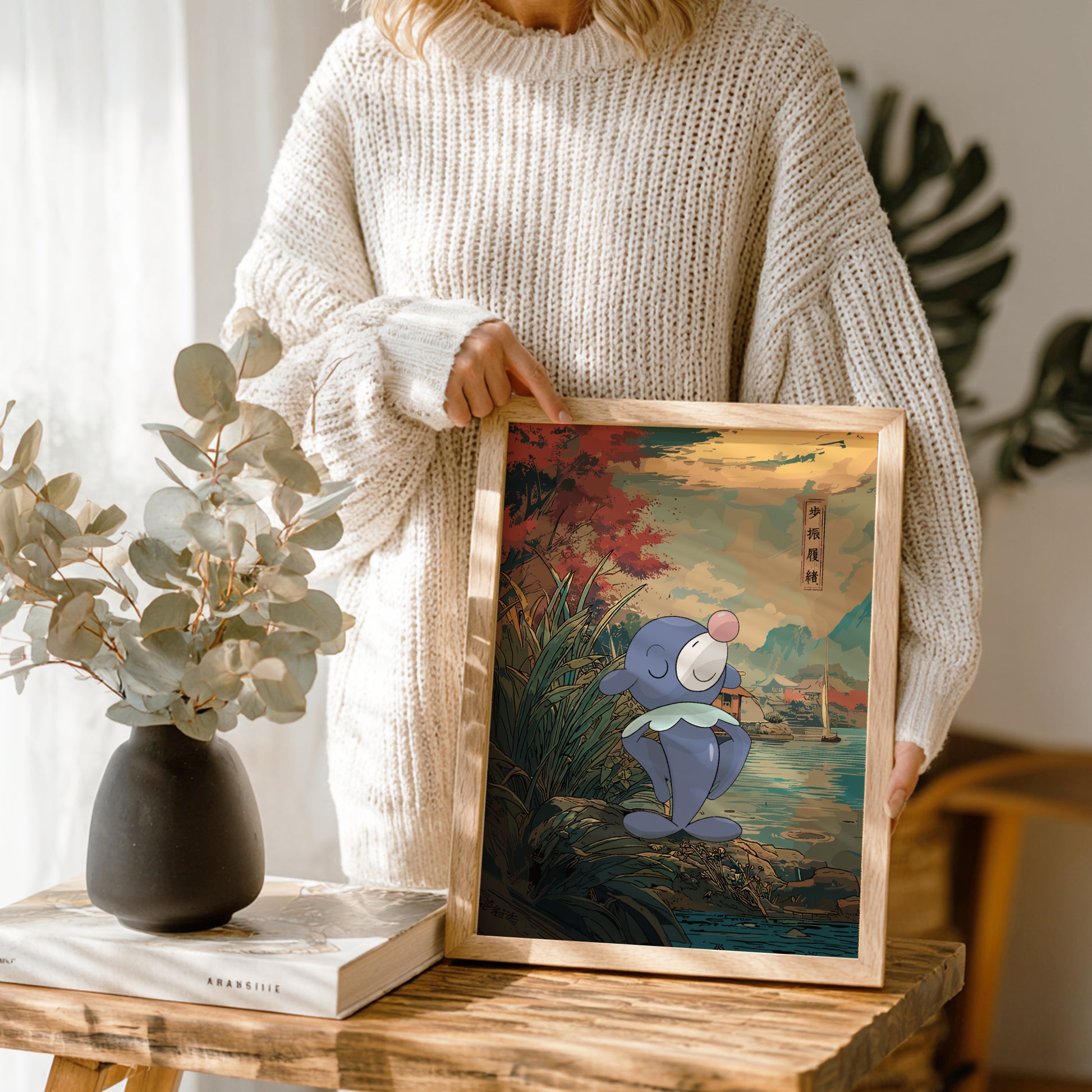 Person holding a framed artwork of Popplio Pokemon character by a vase and book on a wooden table.