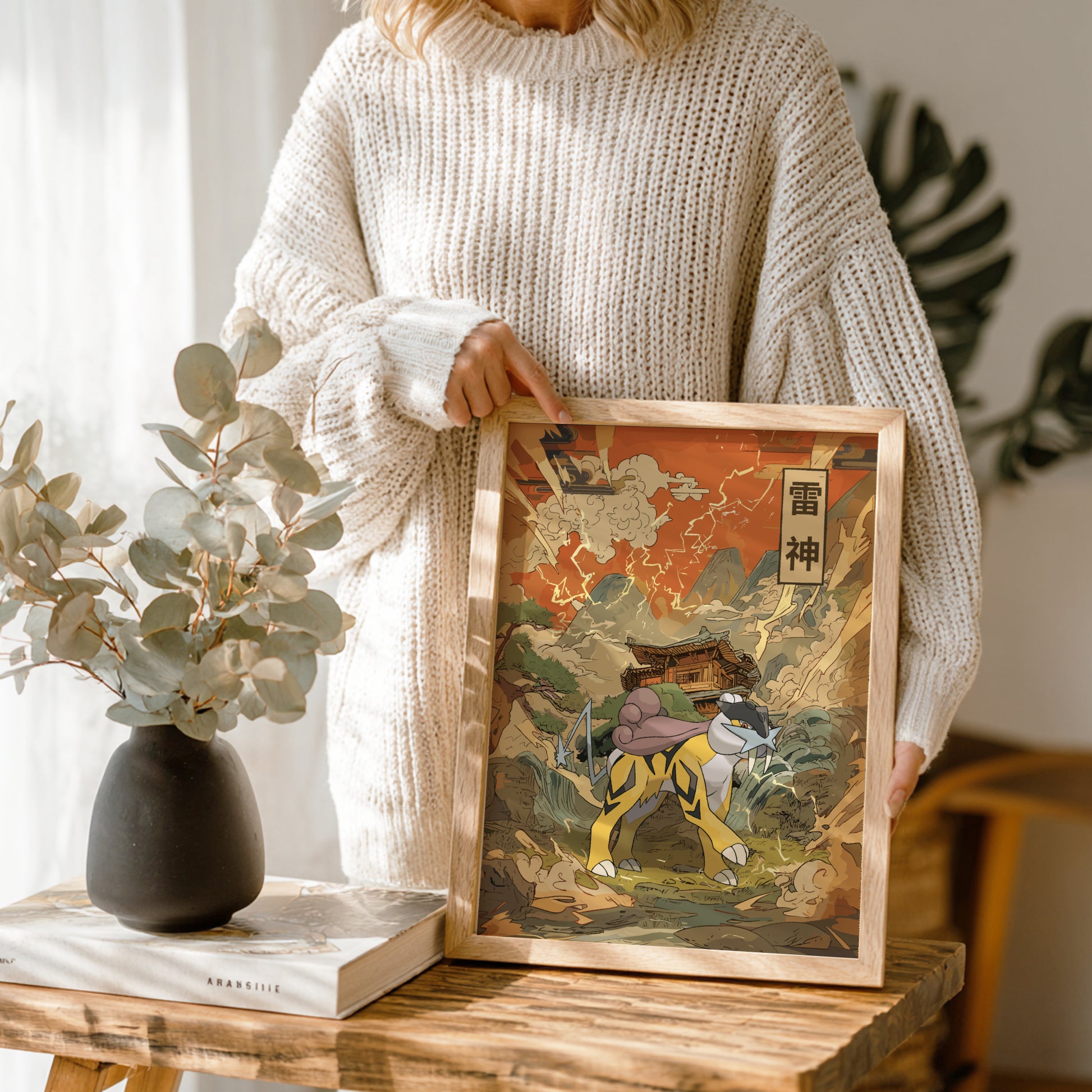 Person holding a framed traditional artwork of Raikou Pokemon on a wooden table with a vase and book in the background.