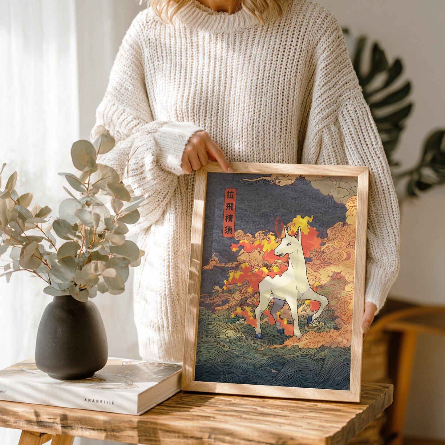 Person holding a framed artwork of Rapidash Pokemon in a colorful landscape on a wooden table.