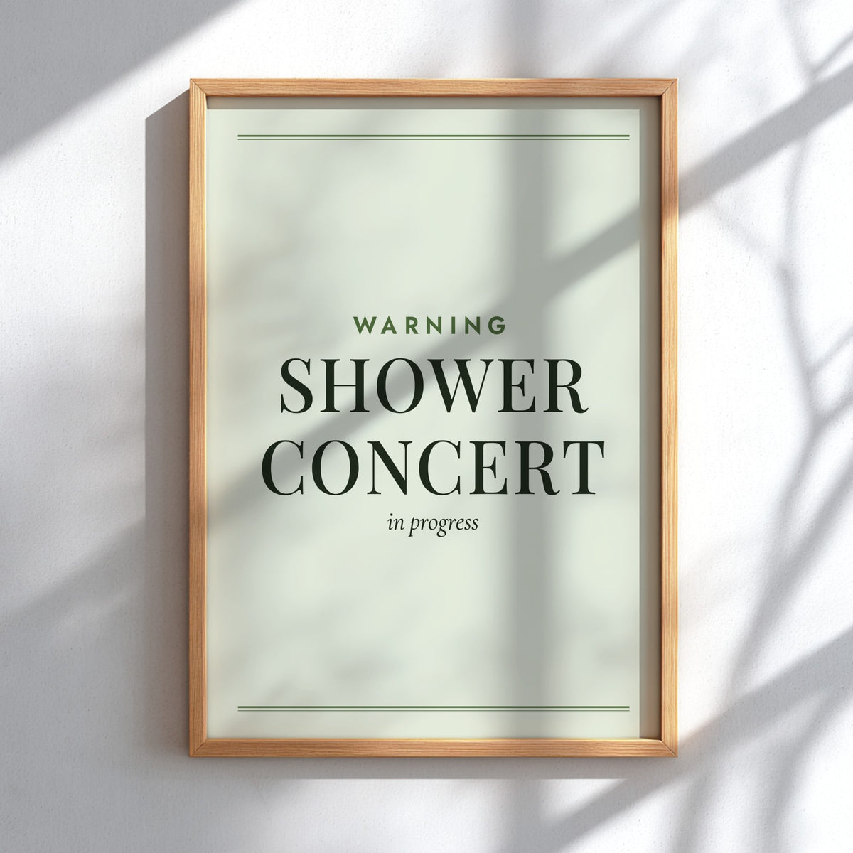 A framed wall art print with the text 'WARNING SHOWER CONCERT in progress' in dark green and black lettering on a pale green background, shown in a light wood frame hanging on a white wall with soft shadow patterns.