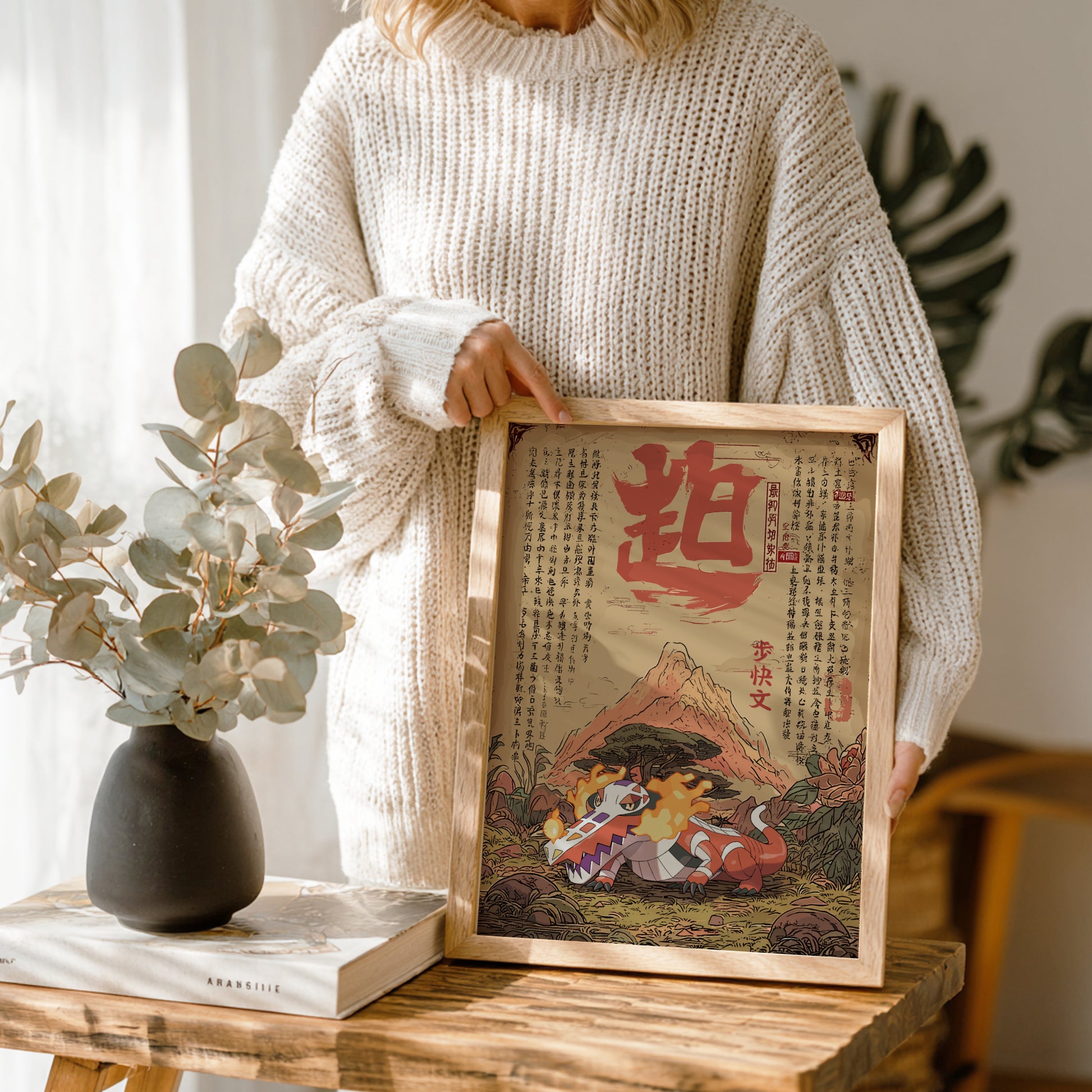 Person holding a framed artwork with a mountain and Skeledirge Pokemon design, standing in a room with plants and a vase.
