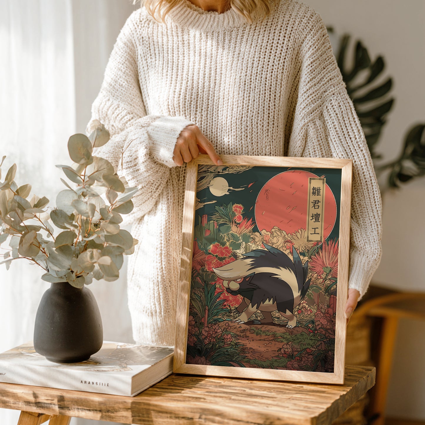 Person holding a framed artwork with a Skuntank Pokemon and red sun, standing next to a vase and books on a wooden table.