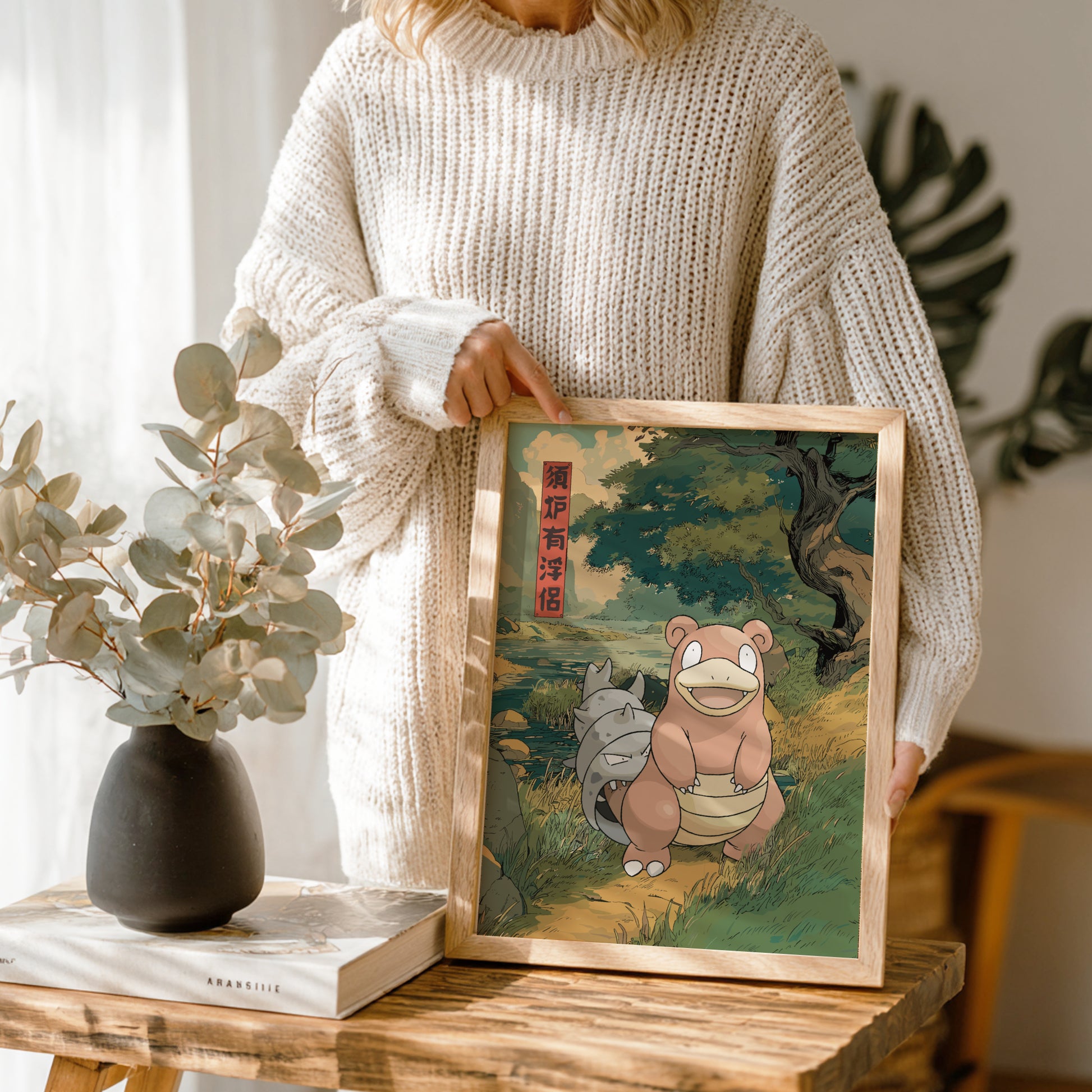 Person holding a framed illustration of Slowbro Pokemon on a wooden table with plants and books in the background.
