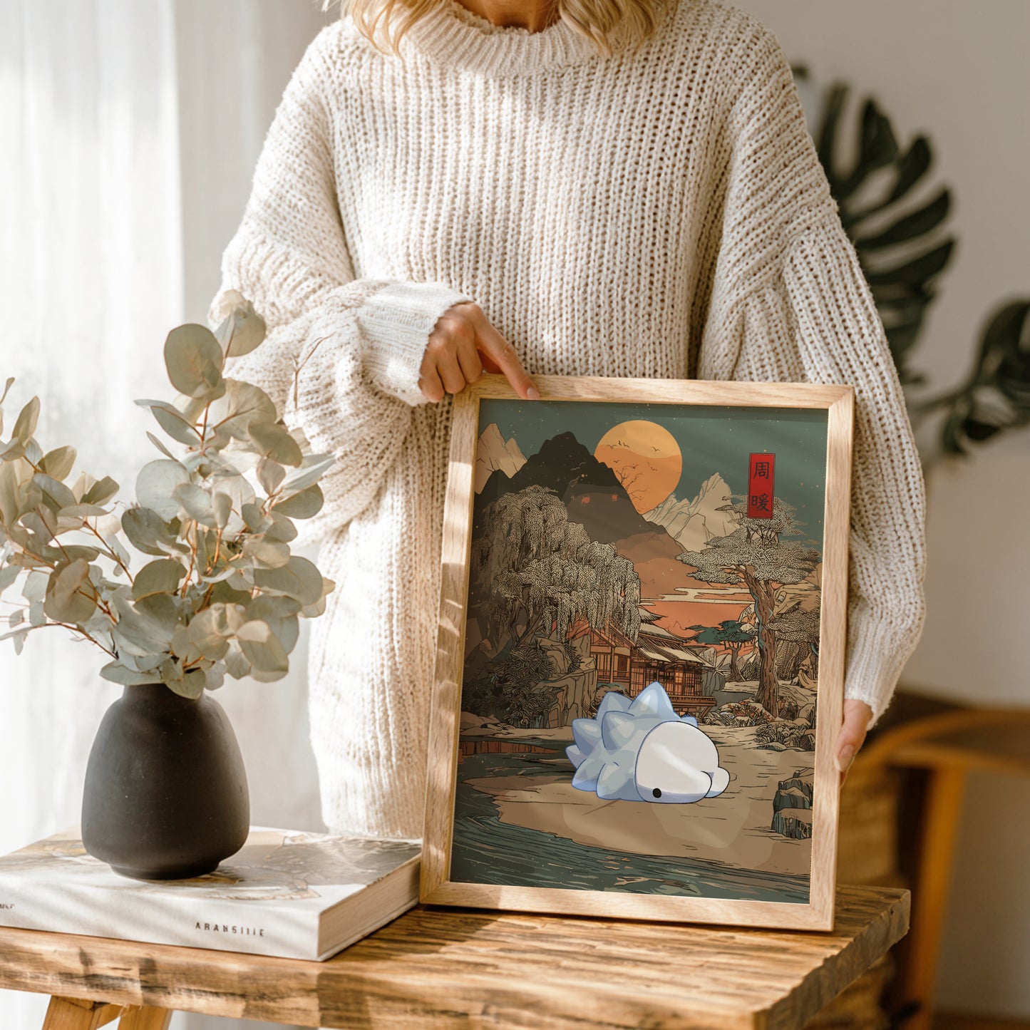 Person holding a framed artwork with a mountain scene and Snom Pokemon on a wooden table.