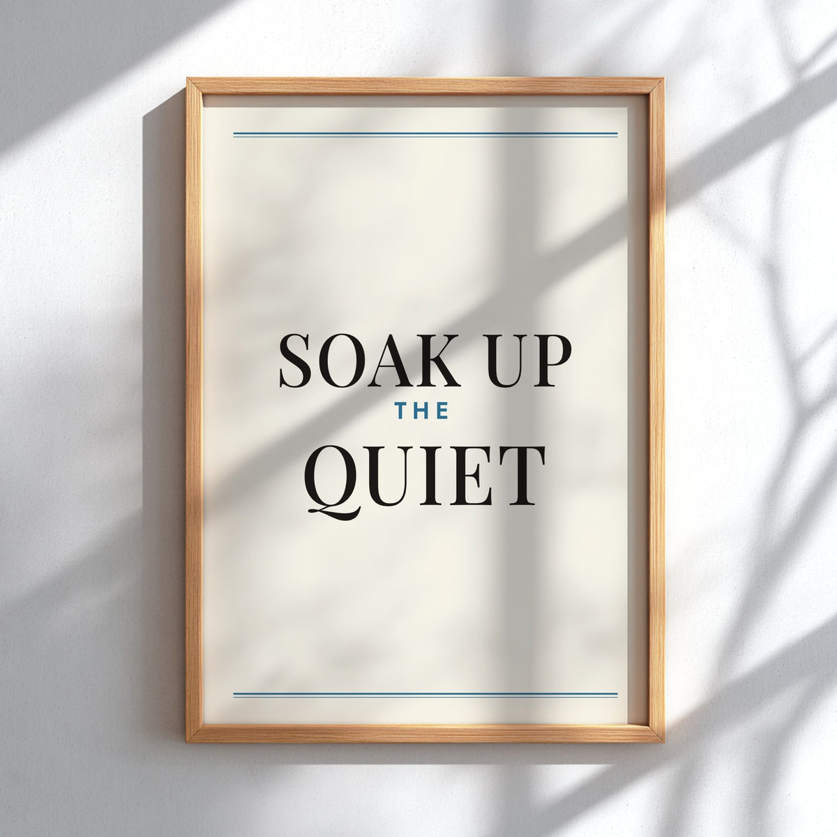 Framed wall art print with the phrase “SOAK UP THE QUIET” in black serif text on an off-white background, with thin blue horizontal lines near the top and bottom and a light natural wood frame, mounted on a white wall with soft shadow patterns.