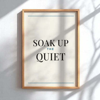 Framed wall art print with the phrase “SOAK UP THE QUIET” in black serif text on an off-white background, with thin blue horizontal lines near the top and bottom and a light natural wood frame, mounted on a white wall with soft shadow patterns.