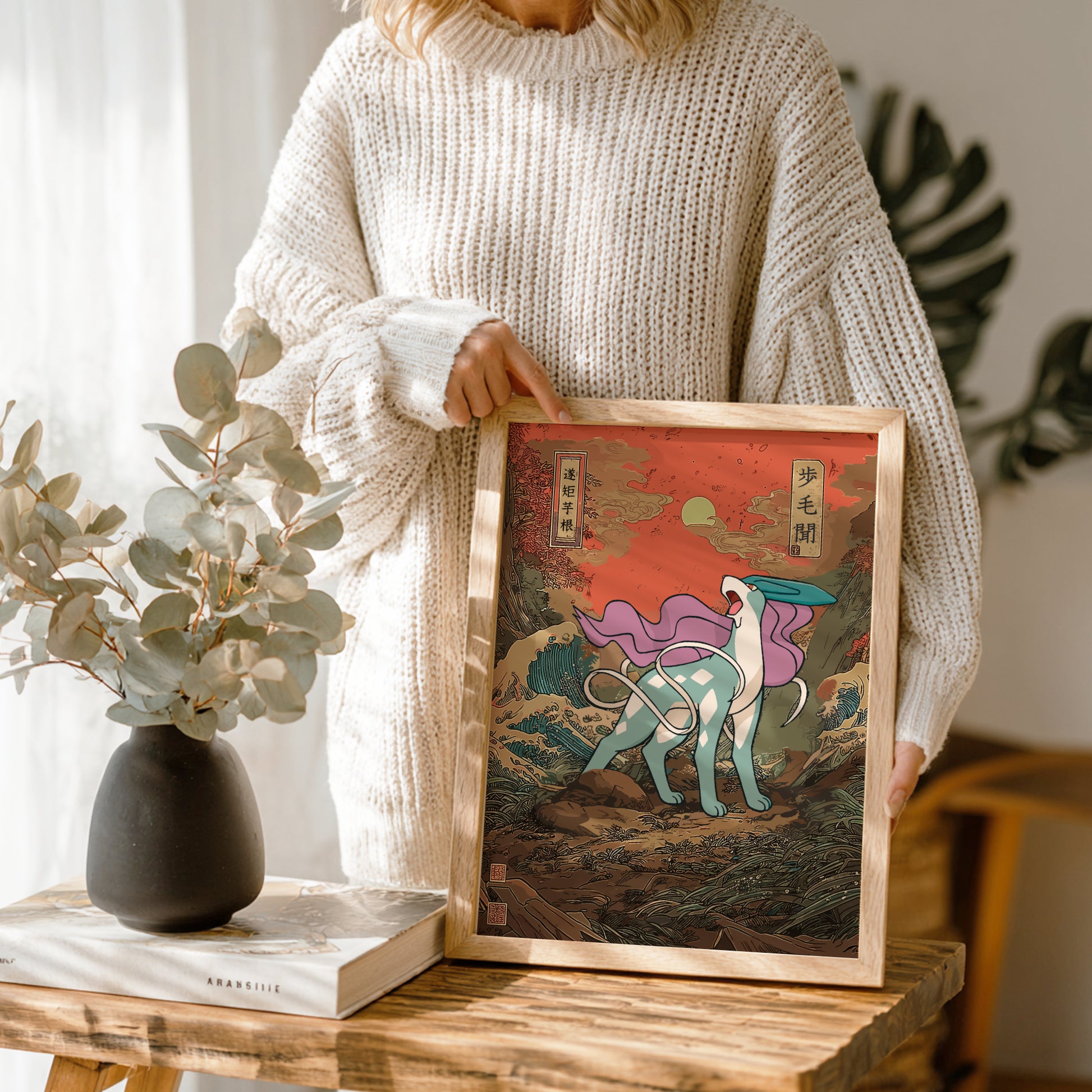 Person holding a framed artwork with a scenic design of Suicune Pokemon, standing next to a vase and book on a wooden table.