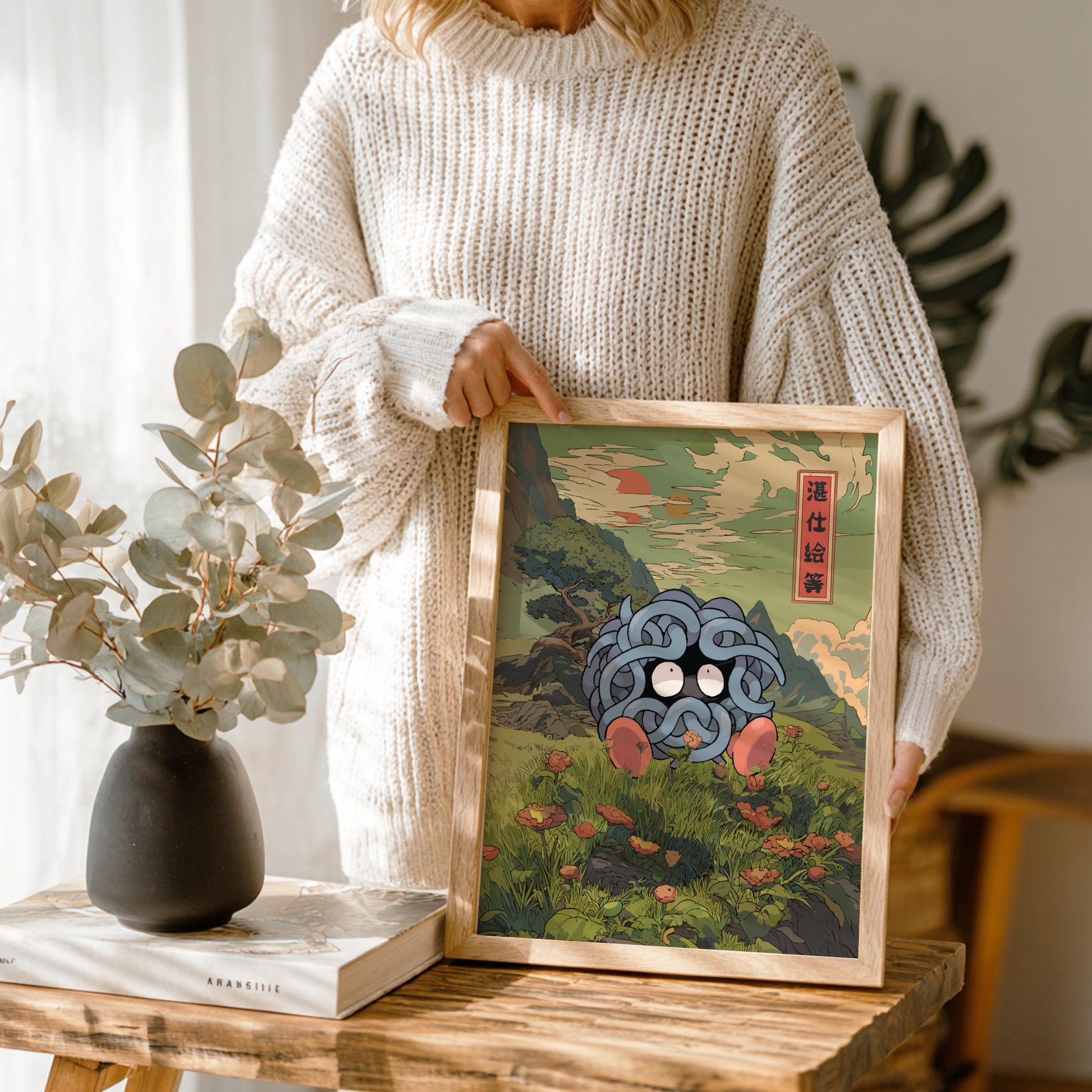 Person holding a framed artwork of Tangela Pokemon with a natural setting and abstract elements, standing in a room with plants and a vase.