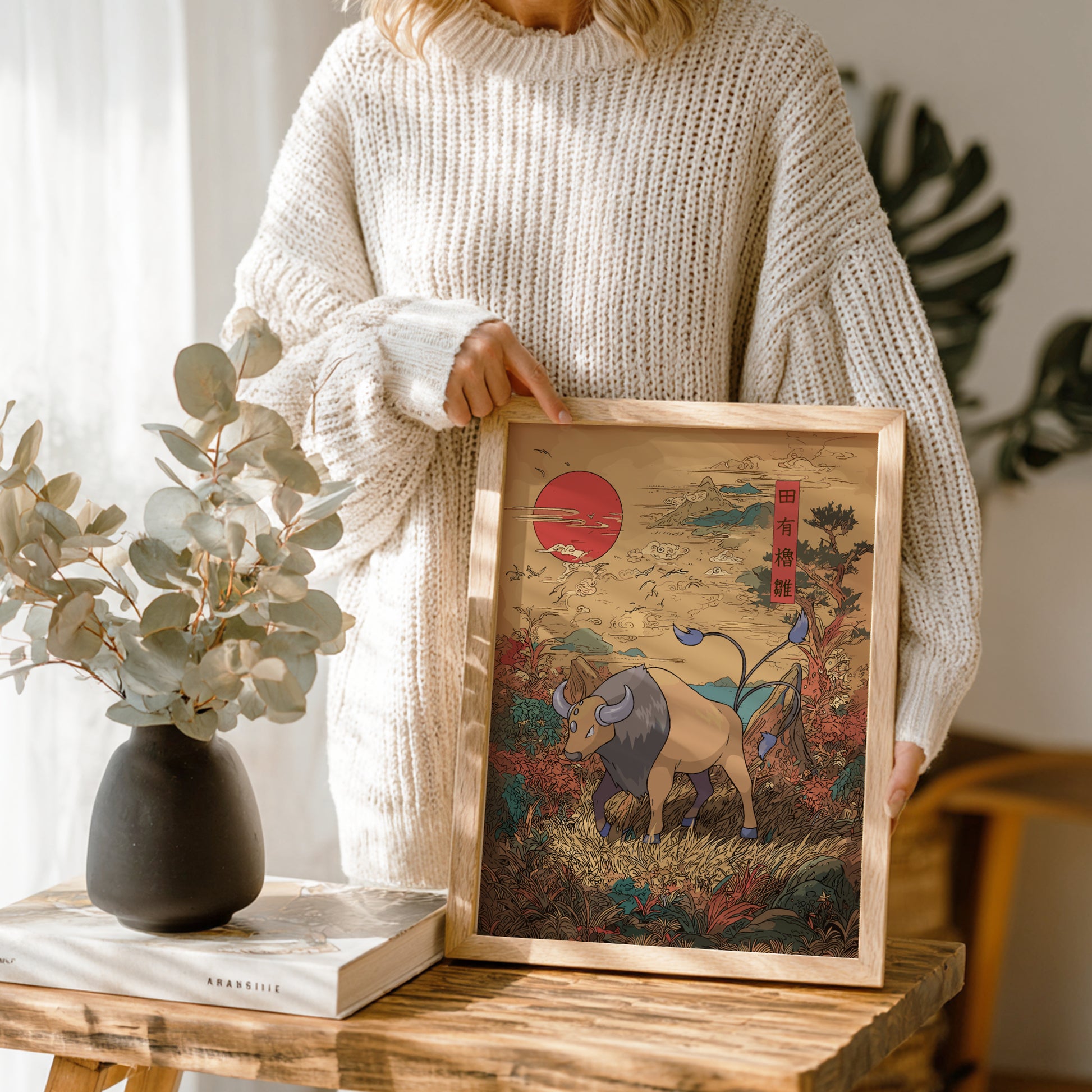 Person holding a framed artwork of Tauros Pokemon in a natural setting, with a vase and book on a wooden table.