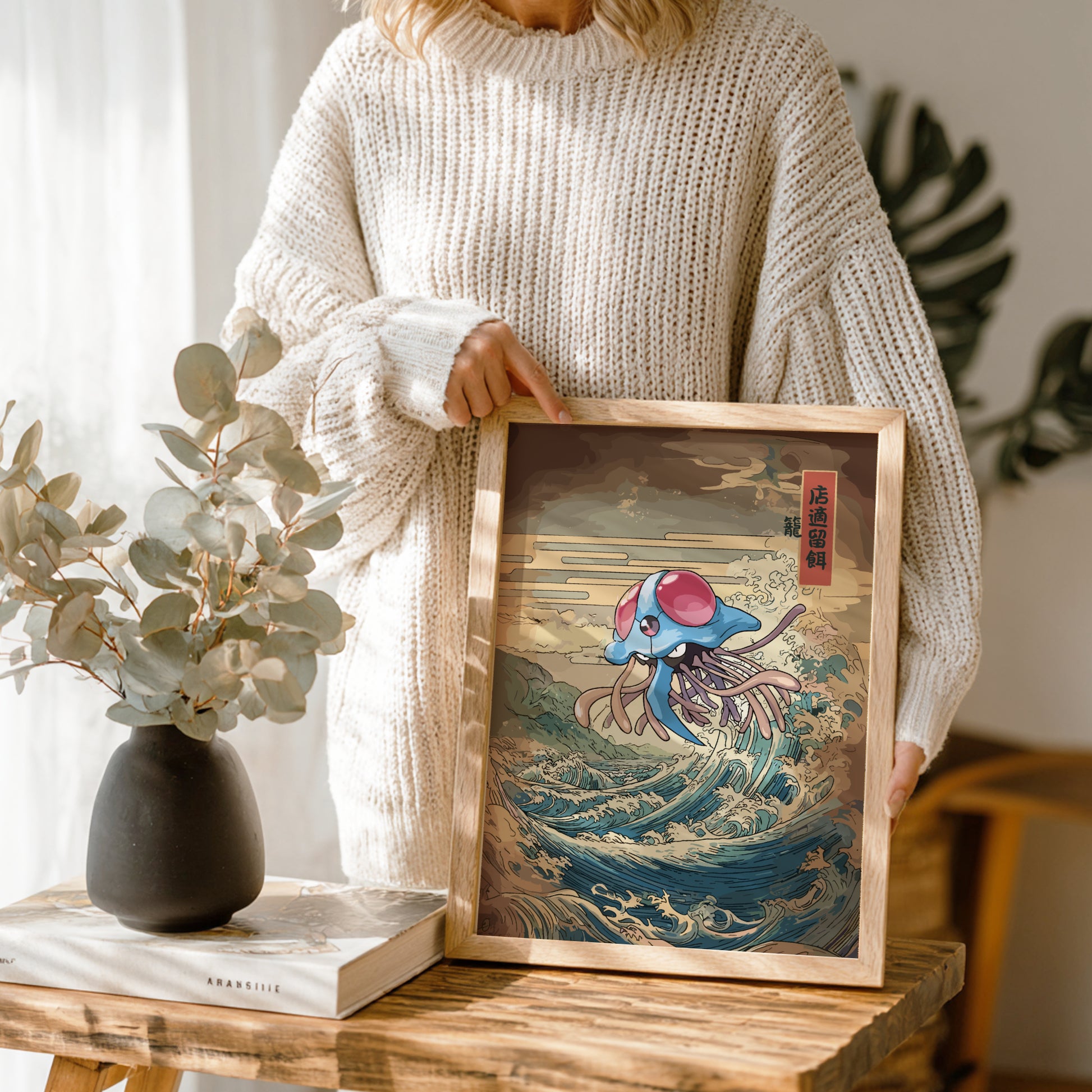 Person holding a framed artwork of Tentacruel Pokemon on a wooden table with a vase and book in the background.
