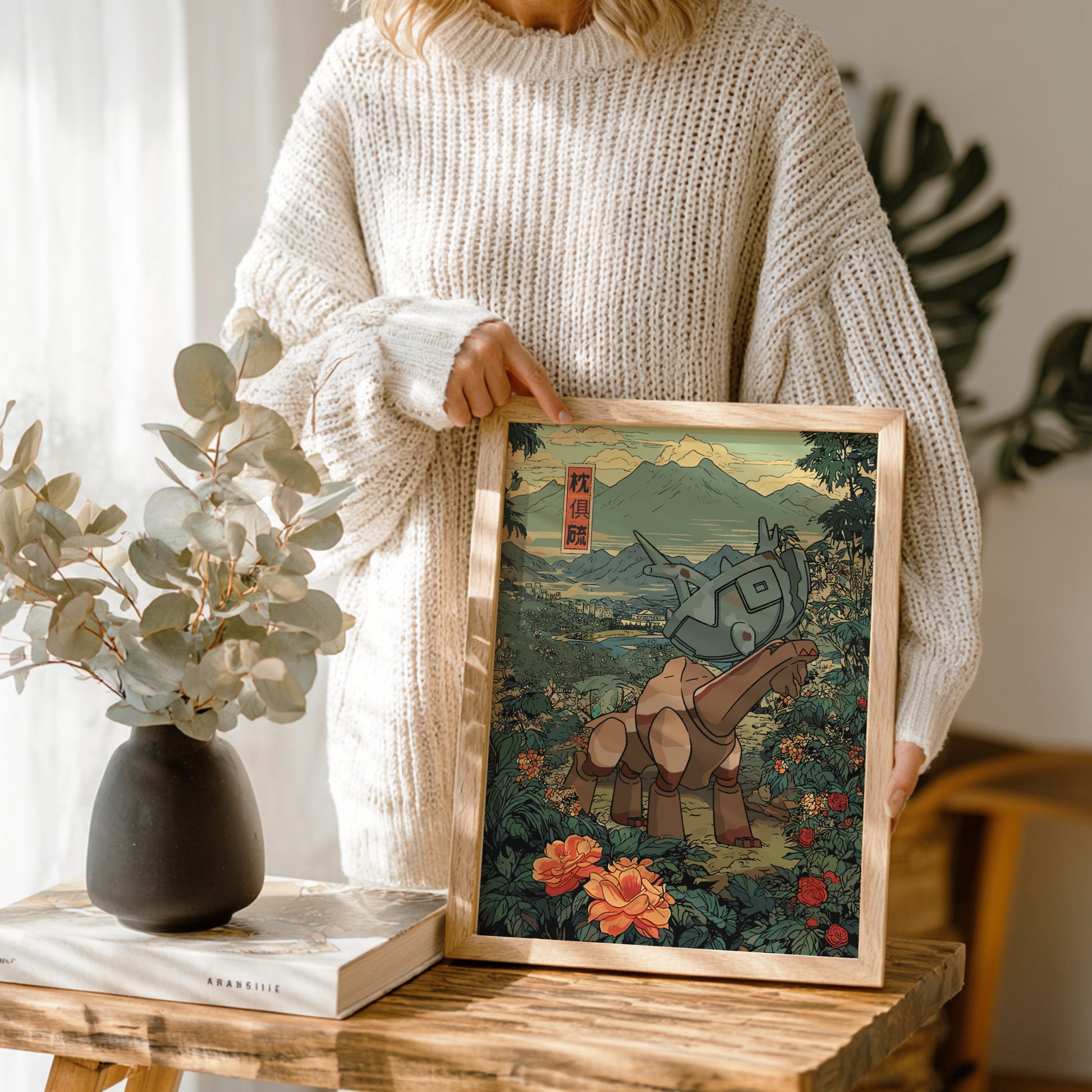 Person holding a framed artwork of a stylized Ting-Lu Pokemon in a natural setting, with a vase and books on a wooden table.