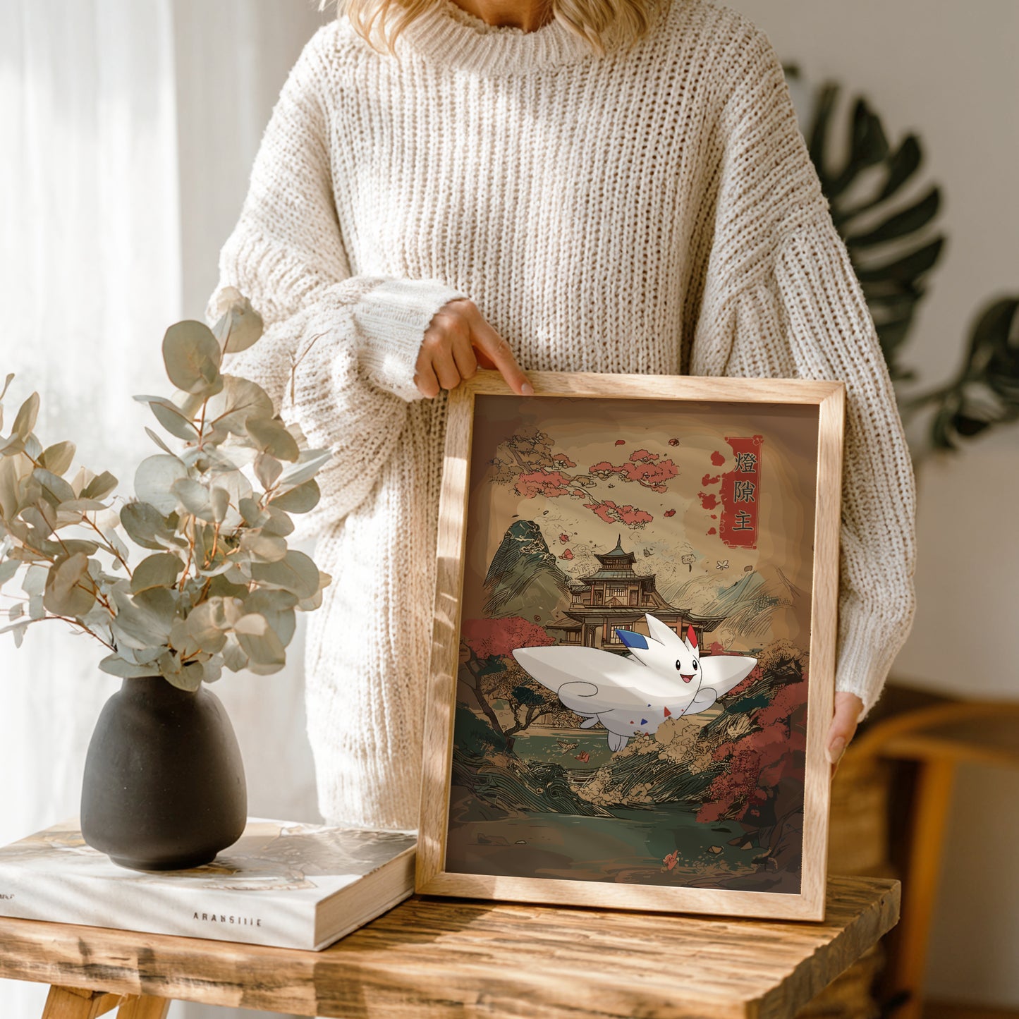 Person holding a framed artwork with a scenic design and Togekiss Pokemon, standing next to a vase and book on a wooden table.