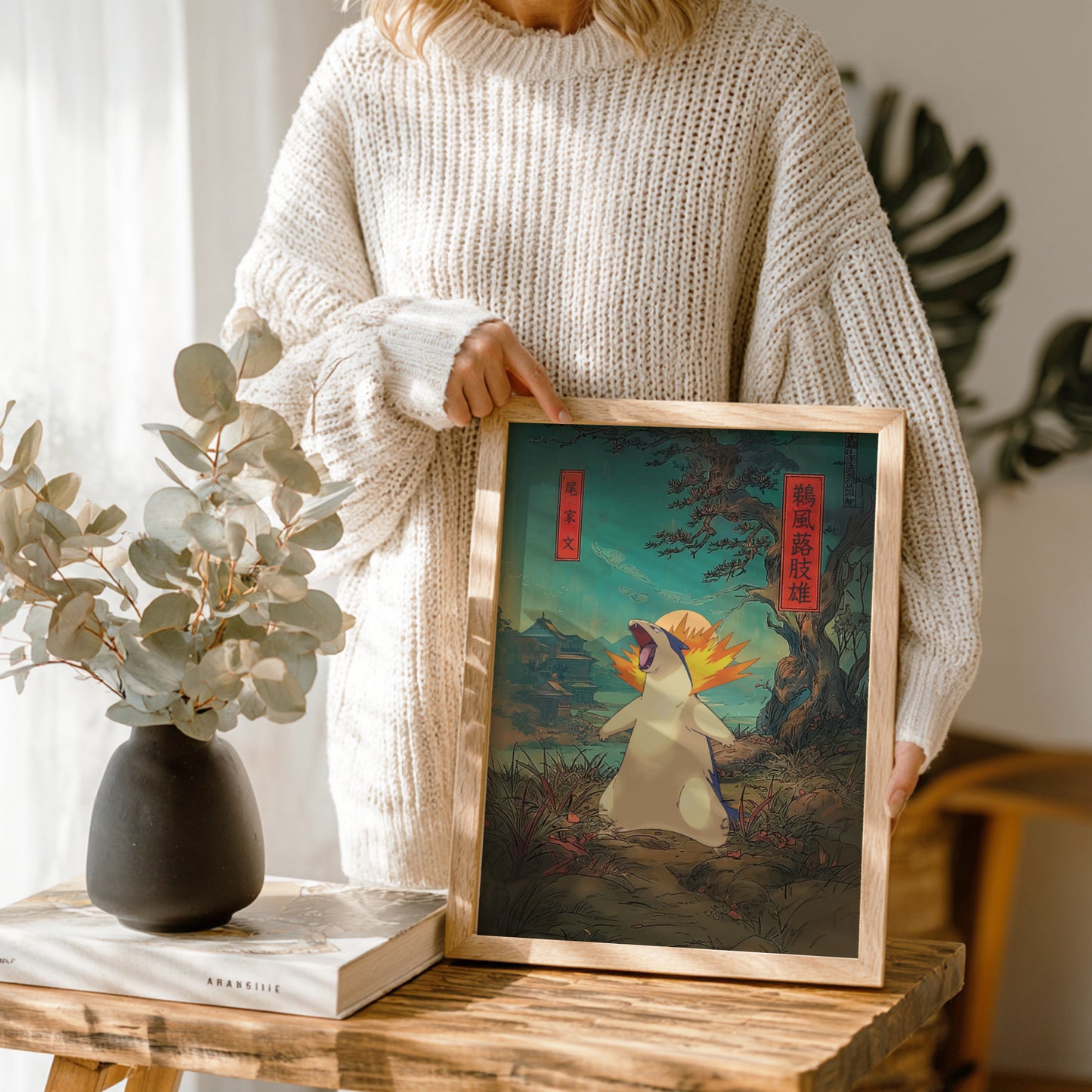 Person holding a framed artwork of Typhlosion Pokemon in a natural setting, with a vase and book on a wooden table.