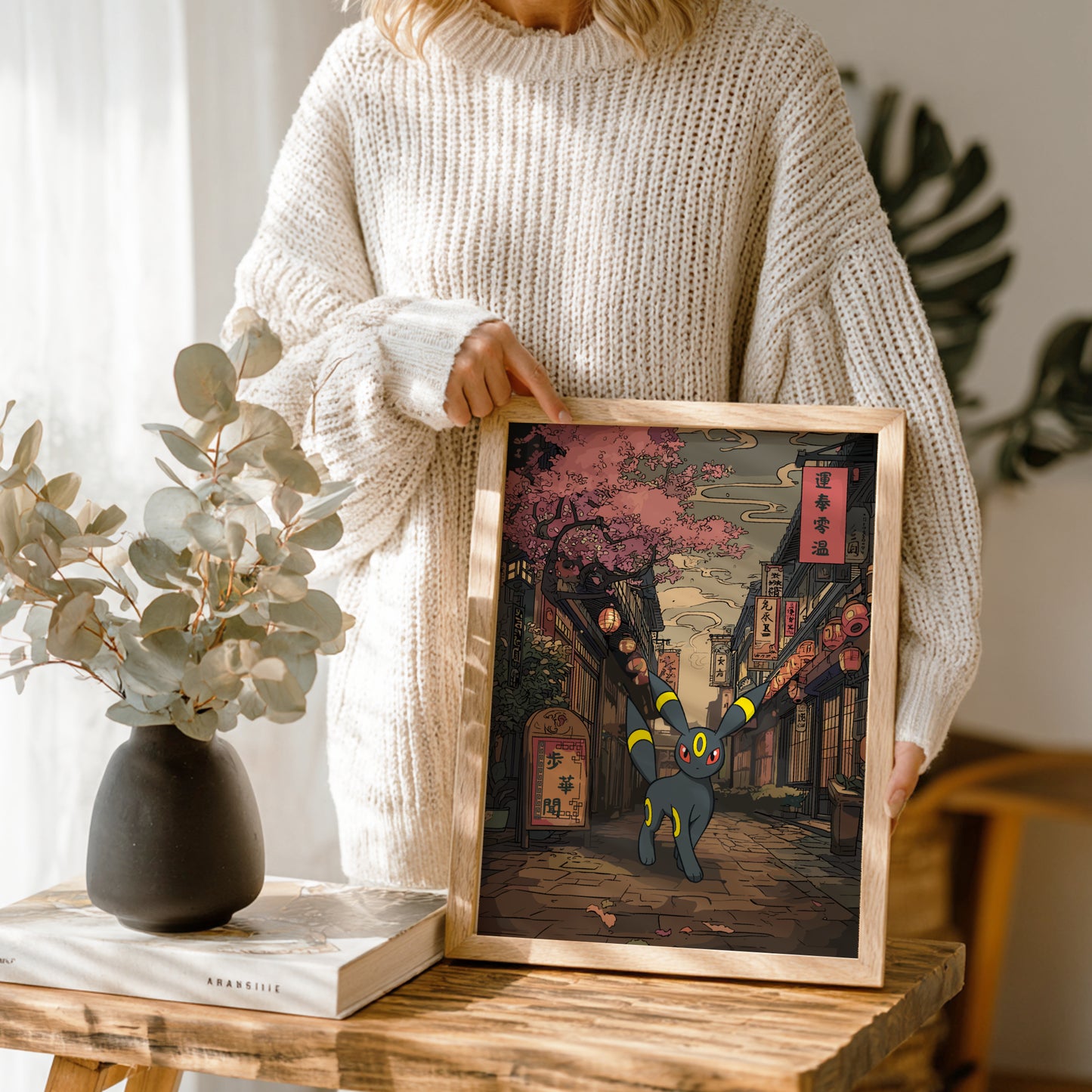 Person holding a framed artwork of Umbreon Pokemon in an urban setting, with a vase and book on a wooden table.