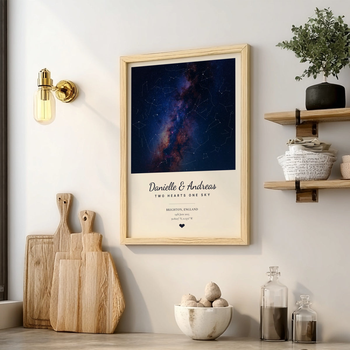 A framed personalized art print featuring a dark blue and purple galaxy night sky with constellations, displayed on a light gray wall in a styled kitchen setting with wooden cutting boards, a shelf, and a plant.
