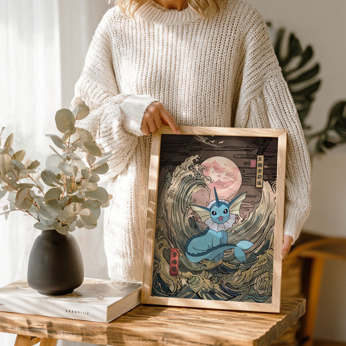 Person holding a framed artwork of Vaporeon Pokemon in a wave, with a vase and book on a wooden table.