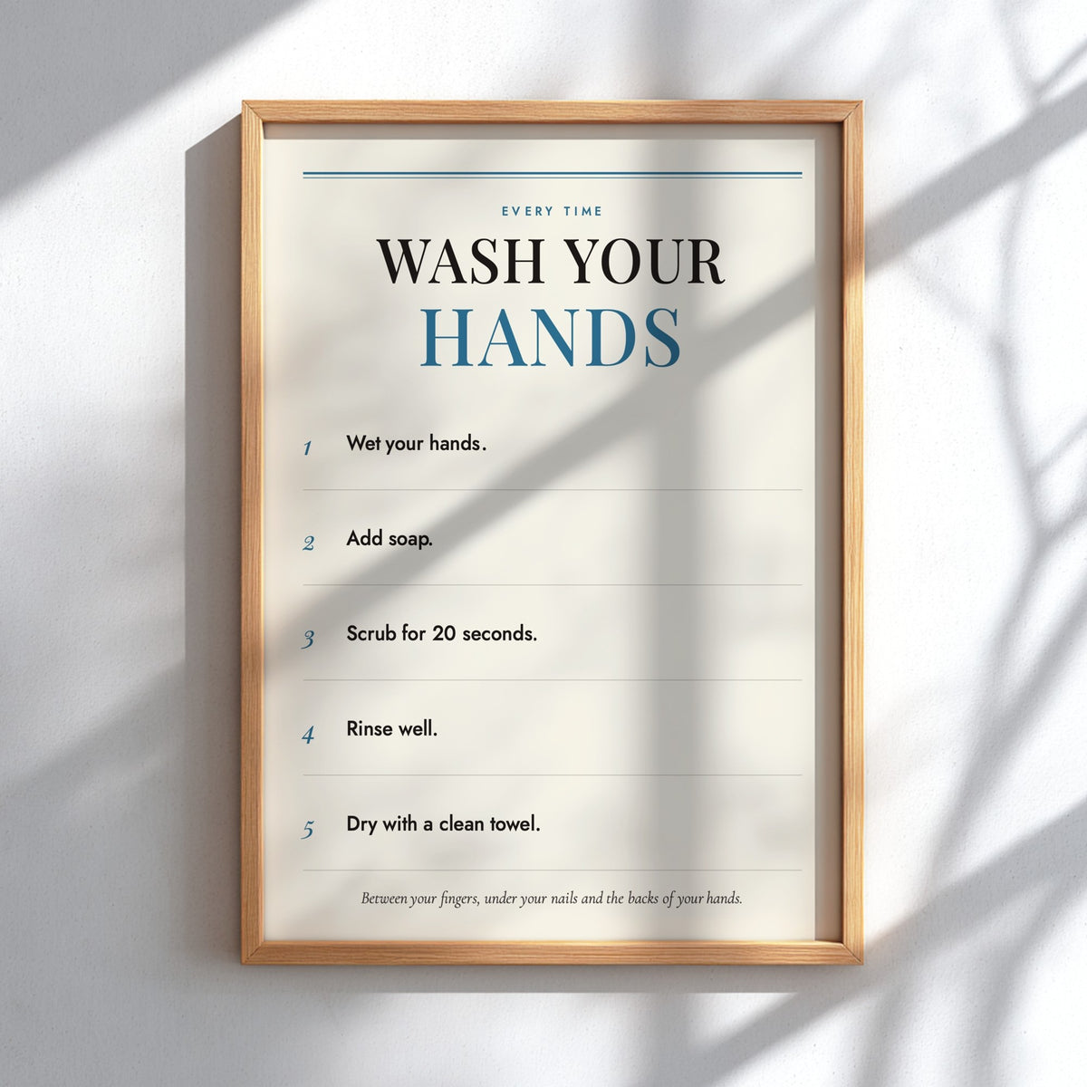 A framed handwashing instruction poster hanging on a white wall, with natural light casting leaf shadows across the surface. The poster reads 'WASH YOUR HANDS' and lists five steps, in a light wood frame.