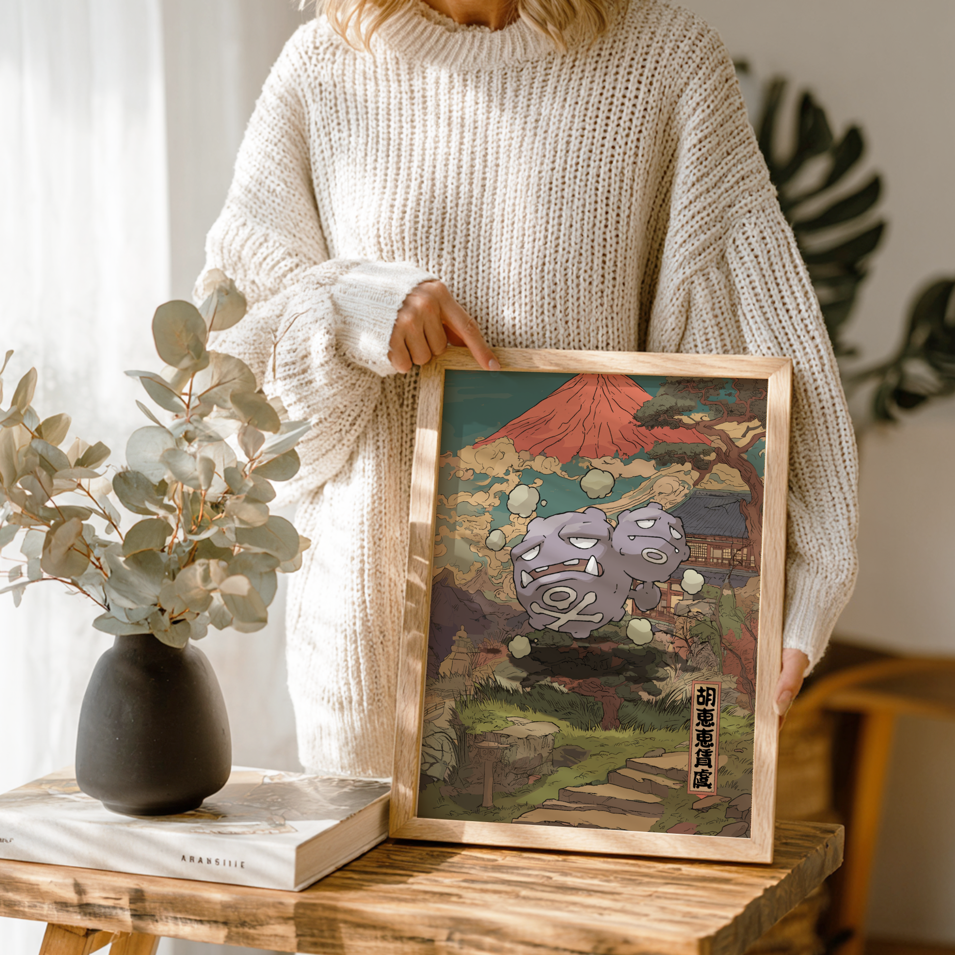 Person holding a framed artwork of Weezing Pokemon with a decorative vase and book on a wooden table.