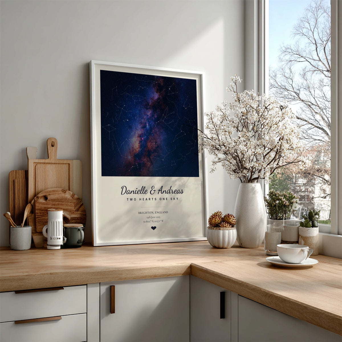 A framed personalized constellation print leaning on a light wood kitchen counter next to a window. The print features a deep blue and purple galaxy design with white text reading 'Danielle & Andreas THE HEARTS GALAXY' and 'MADE FOR LONDON, 2024'.