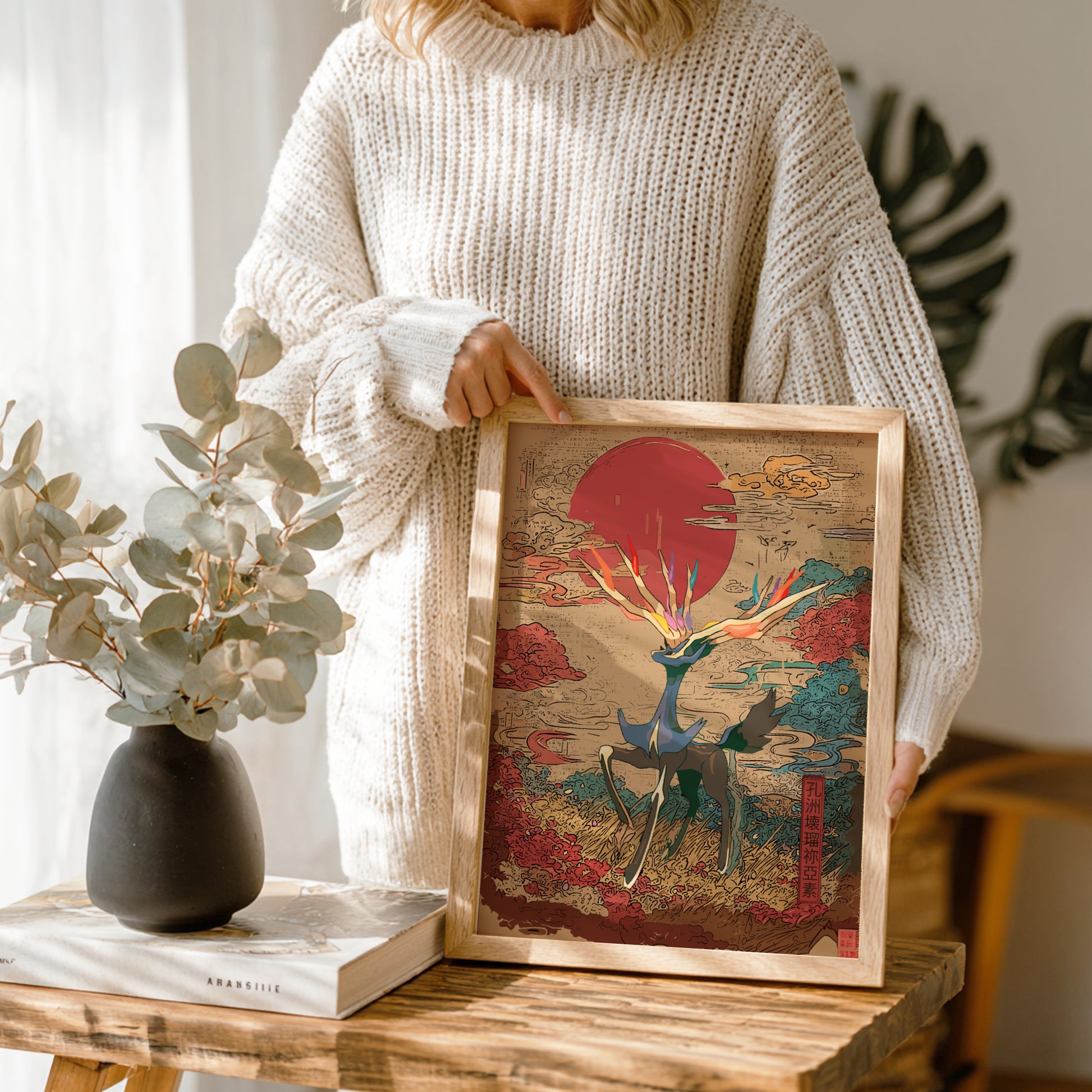 Person holding a framed artwork with Xerneas Pokemon and red sun design, standing in a room with plants and a vase.