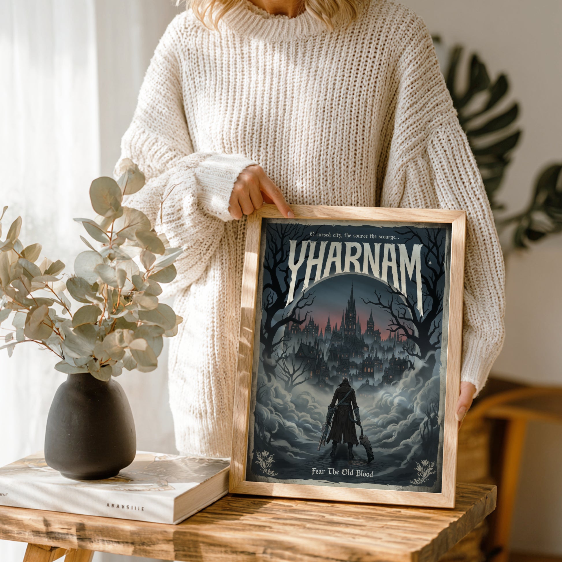 Person holding a framed poster with 'Yharnam' design, standing next to a vase and book on a wooden table.