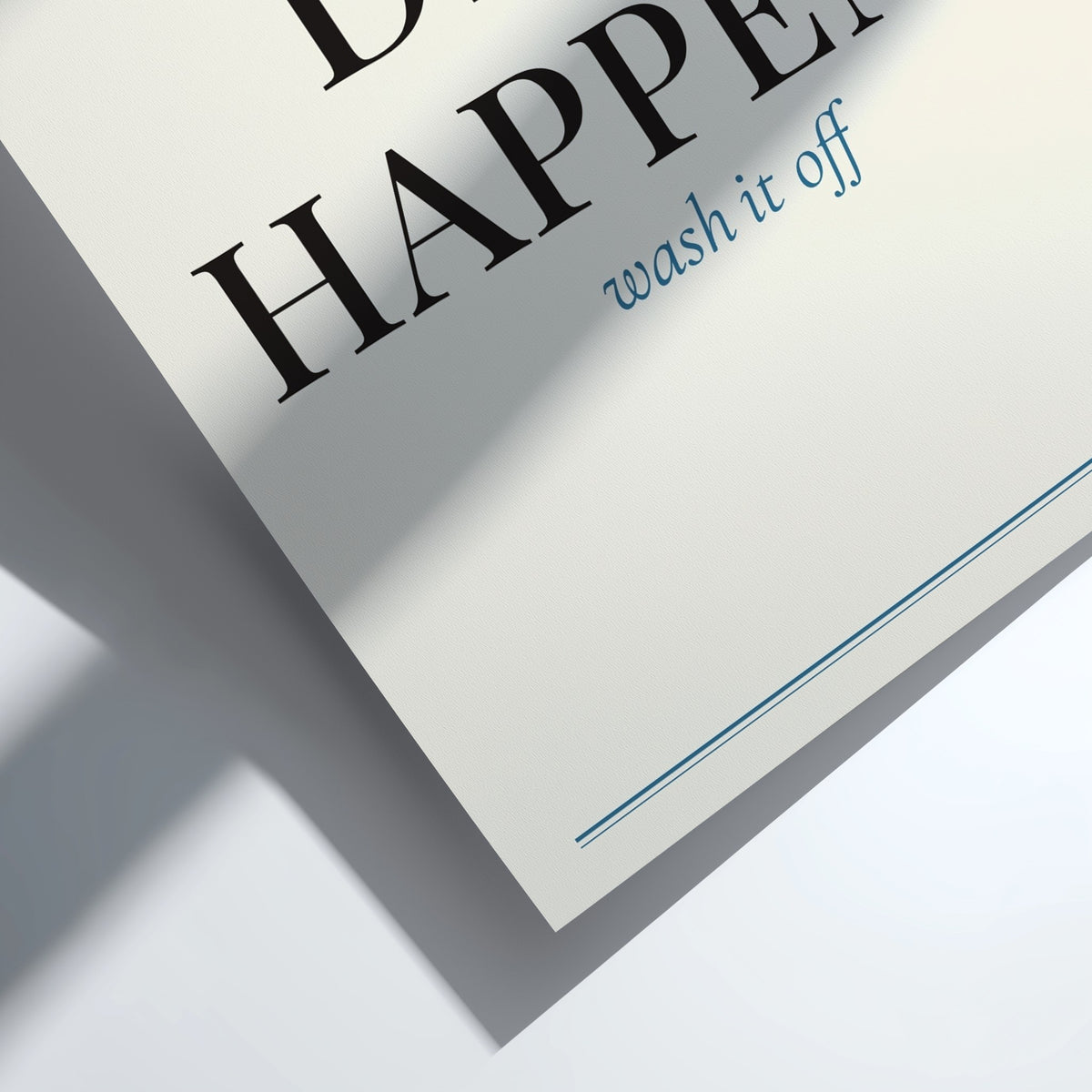 A close-up angled shot of a white paper label or tag printed with the words 'HAPPEN wash it off' in black and blue text, casting a soft shadow on a white surface.