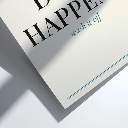A close-up angled shot of a white paper label or tag printed with the words 'HAPPEN wash it off' in black and blue text, casting a soft shadow on a white surface.