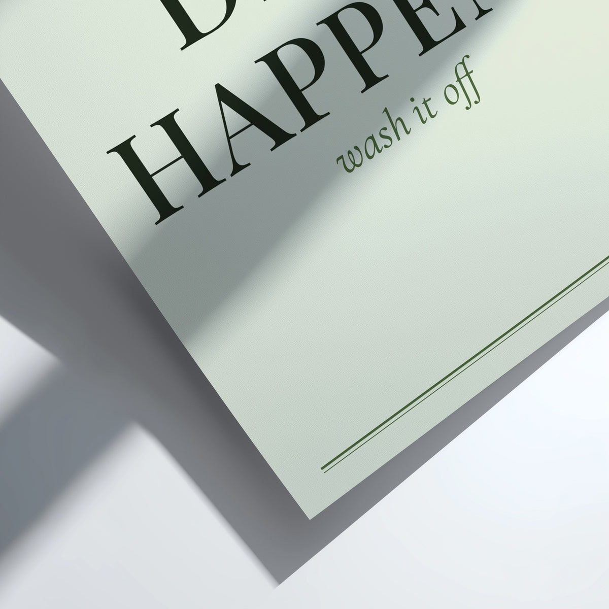 A close-up of a pale sage green paper card or label with black serif text reading 'HAPPEN' and smaller text 'wash it off', shown on a white surface with a soft shadow.