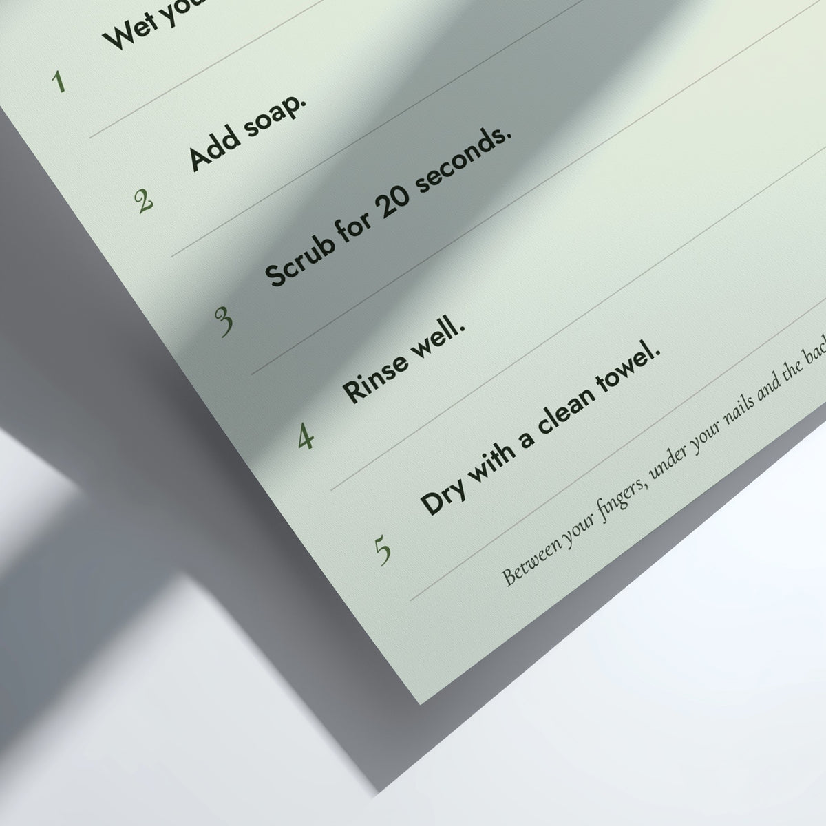A close-up of a printed step-by-step instruction sheet showing how to wash or clean something, with numbered steps from 1 to 5 in dark green text on a pale mint background. The sheet is photographed at an angle on a white surface with soft shadows.