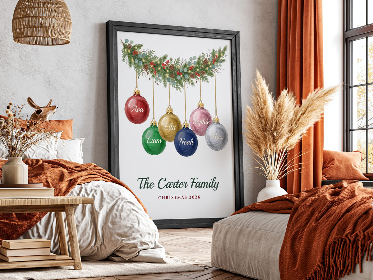 A framed personalized Christmas print leaning against a wall in a styled bedroom. The print shows a green garland with colorful hanging ornaments and the text 'The Carter Family Christmas 2026'.