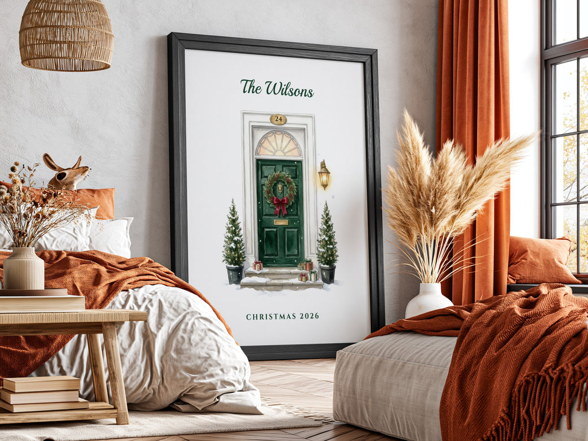 A framed personalized Christmas print leaning against a wall in a cozy bedroom. The black-framed poster features a green front door with a wreath, small Christmas trees, and the text 'The Wilsons' at the top and 'Christmas 2026' at the bottom.