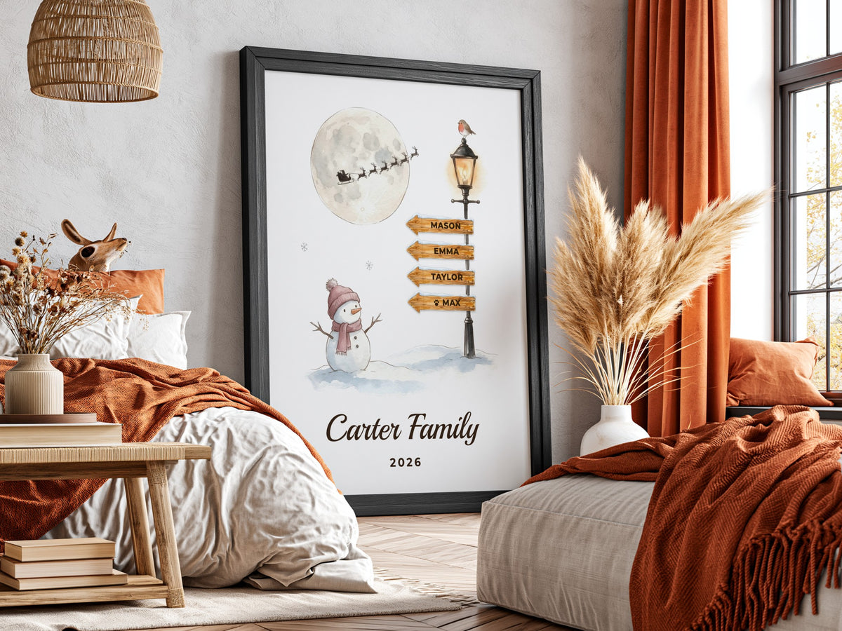 A framed personalized art print leaning against a wall in a cozy bedroom setting. The print features a winter scene with a snowman, a full moon, a streetlamp, and wooden signposts, with the text 'Carter Family 2026' at the bottom.