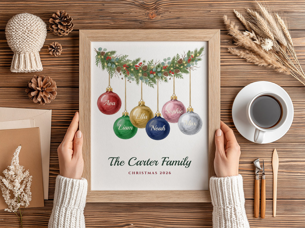 A framed personalized Christmas print held by two hands over a rustic wooden table. The print features a watercolor green garland with colorful hanging ornaments labeled with family names and the text 'The Carter Family Christmas 2026'.