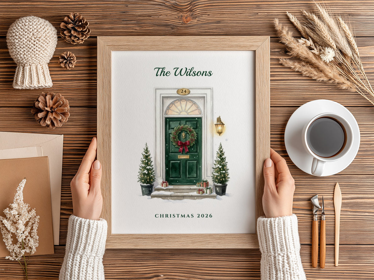 A framed personalized Christmas print held by two hands over a wooden table. The print features an illustration of a green front door with a wreath and the text 'The Wilsons' and 'Christmas 2026'.