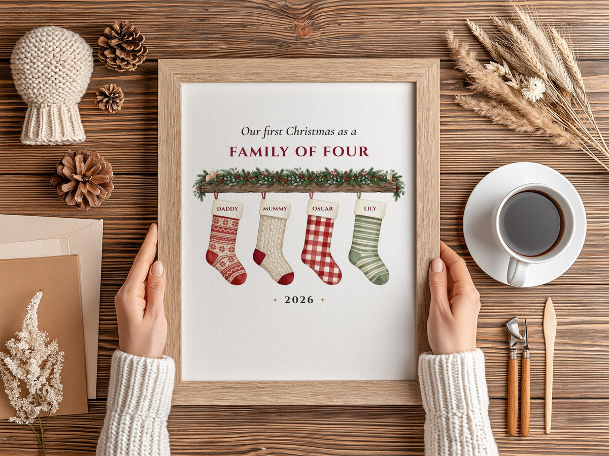 A framed baby milestone chart held by two hands over a wooden table, surrounded by cozy flat-lay props including a cup of coffee, pinecones, dried grass, and a knitted hat.