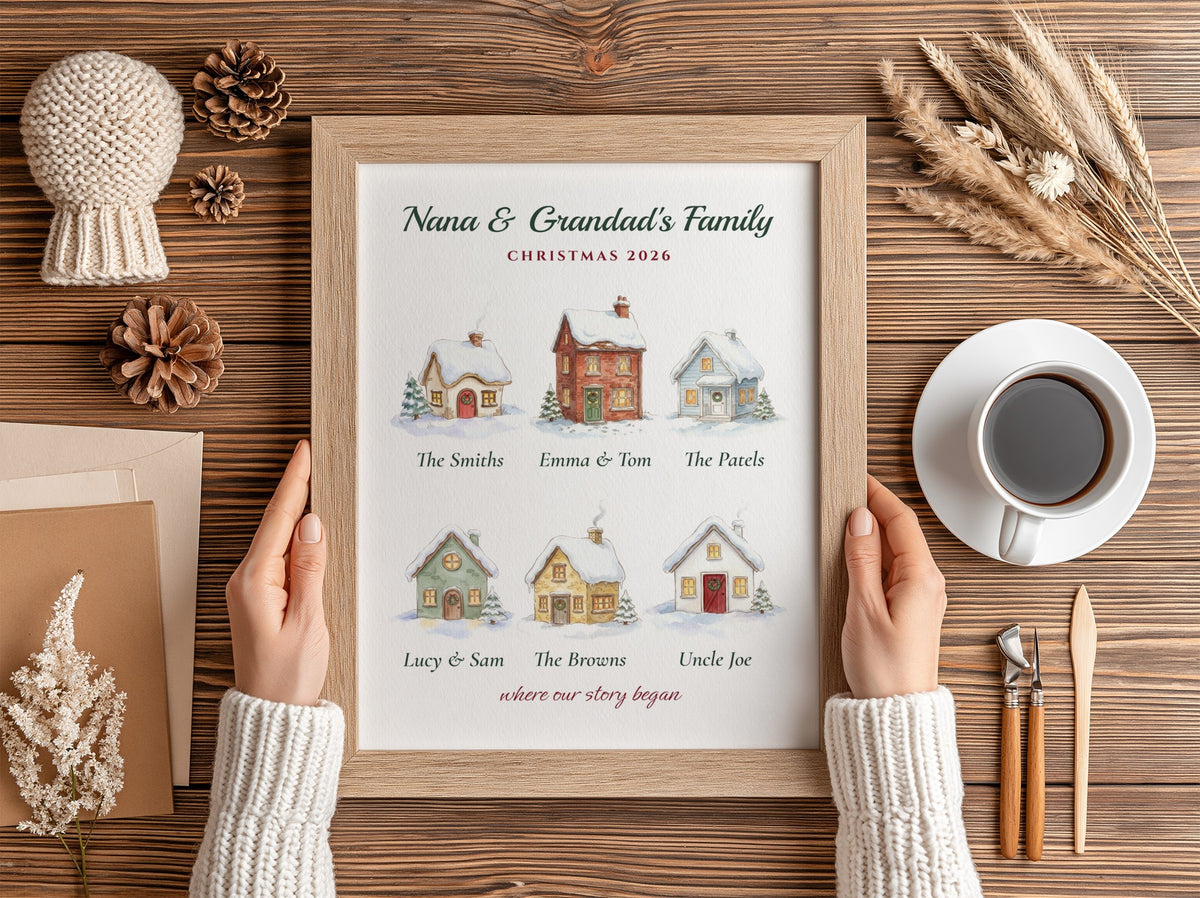 A personalized framed Christmas print held by two hands over a wooden table. The print features watercolor illustrations of holiday houses with custom family names underneath, surrounded by cozy flat-lay props including a cup of coffee, pinecones, dried grass, and a knit hat.