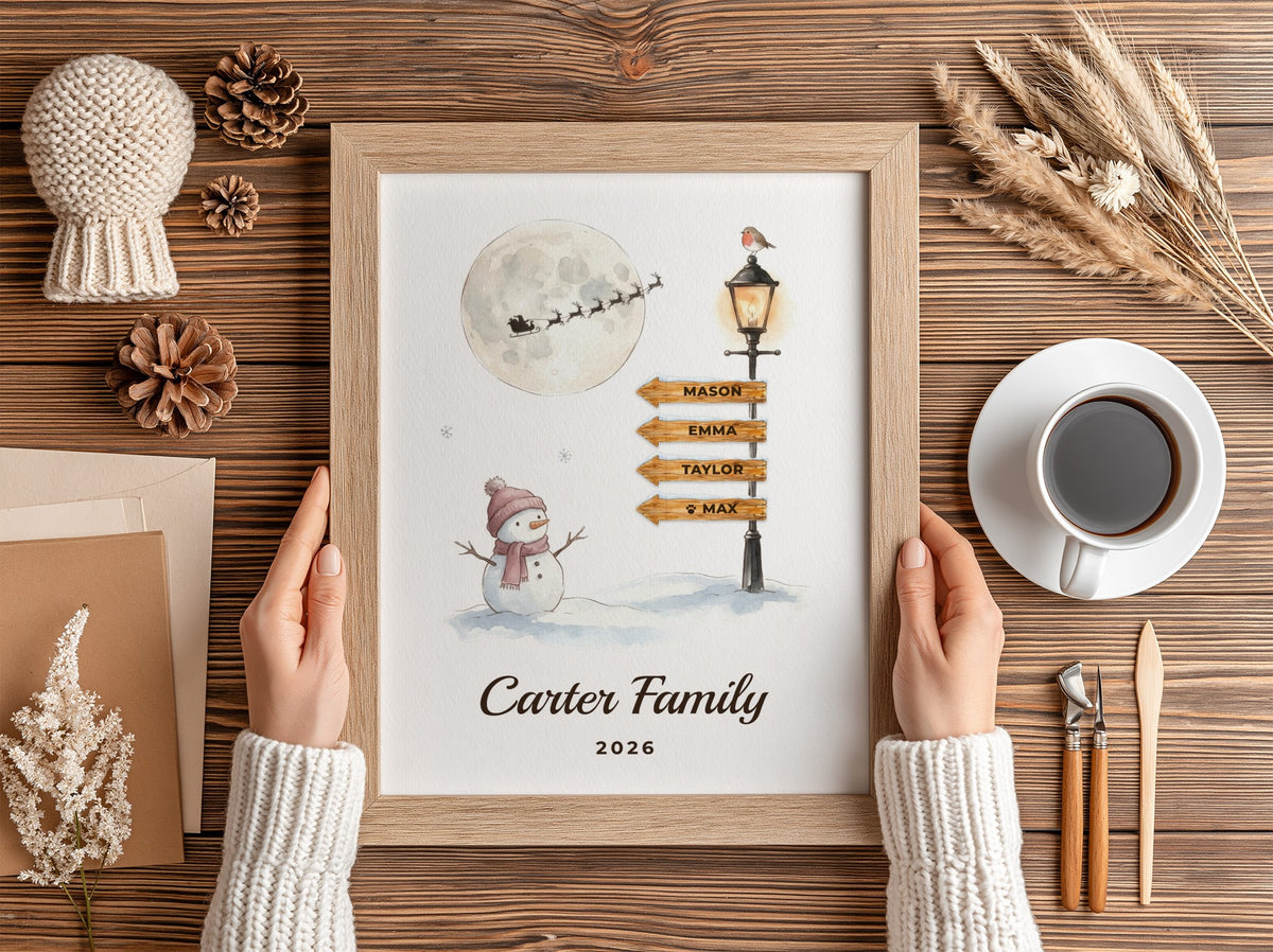 A framed personalized Christmas print held by two hands over a wooden table. The print features a watercolor winter scene with a snowman, moon, sleigh, and directional street signs reading 'MARC', 'EMILY', 'TAYLOR', 'Y MA', with 'Carter Family 2026' written below.