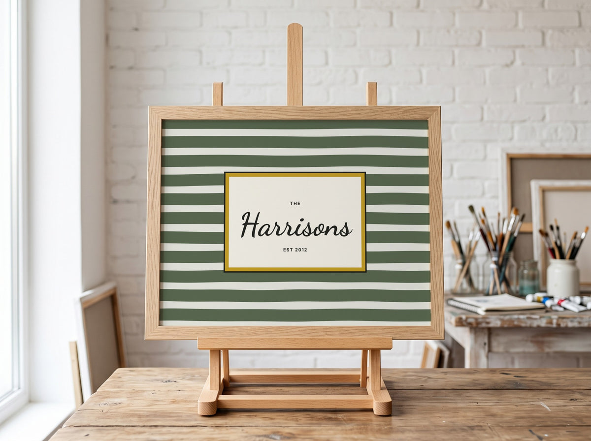 A personalized framed art print with green and white horizontal stripes, featuring a central cream label with the name 'The Harrisons Est 2012' in black script, displayed on a wooden easel in a bright, modern art studio.