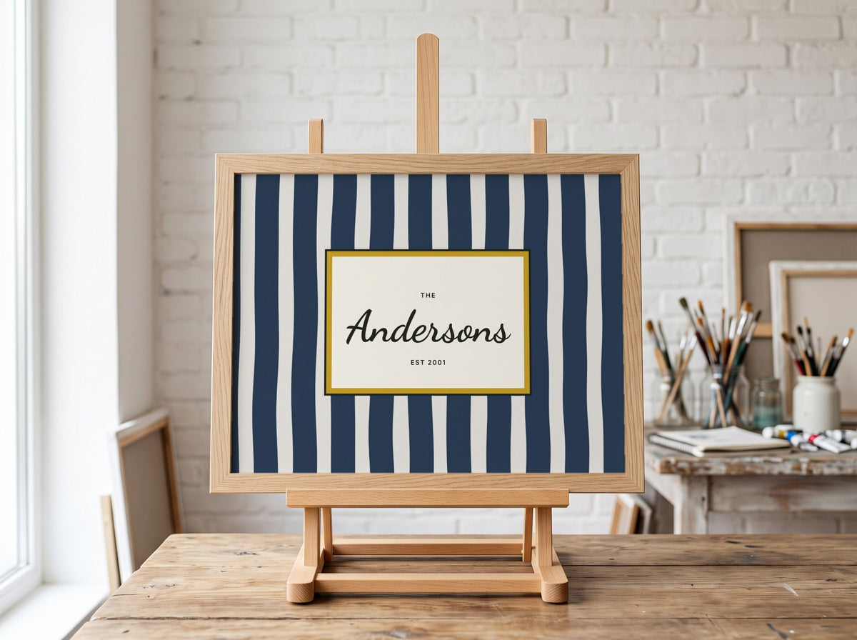 A personalized framed sign with navy and white vertical stripes and a cream center panel reading 'The Andersons EST 2021', displayed on a wooden easel in a bright, styled room with a white brick wall and art supplies.