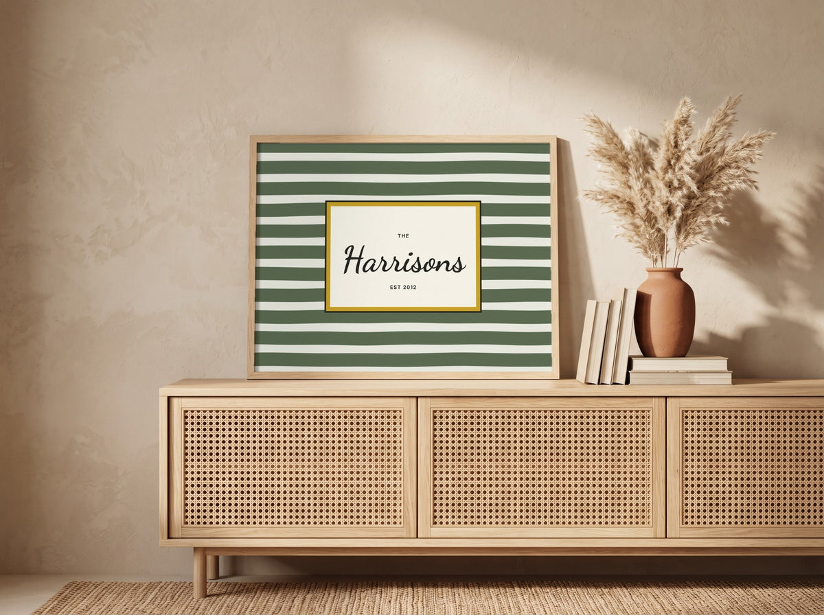 A framed personalized art print with green and white horizontal stripes and a gold border, leaning on a light wood credenza in a styled room setting next to a vase of pampas grass.