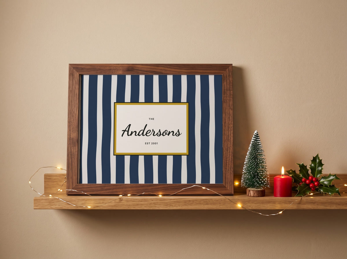 A framed personalized art print with navy and white vertical stripes, featuring a custom name 'Anderson' in black script on a cream background with a gold border. The frame is a medium brown wood, styled on a wooden shelf next to a small Christmas tree, a red candle, and holly against a beige wall.
