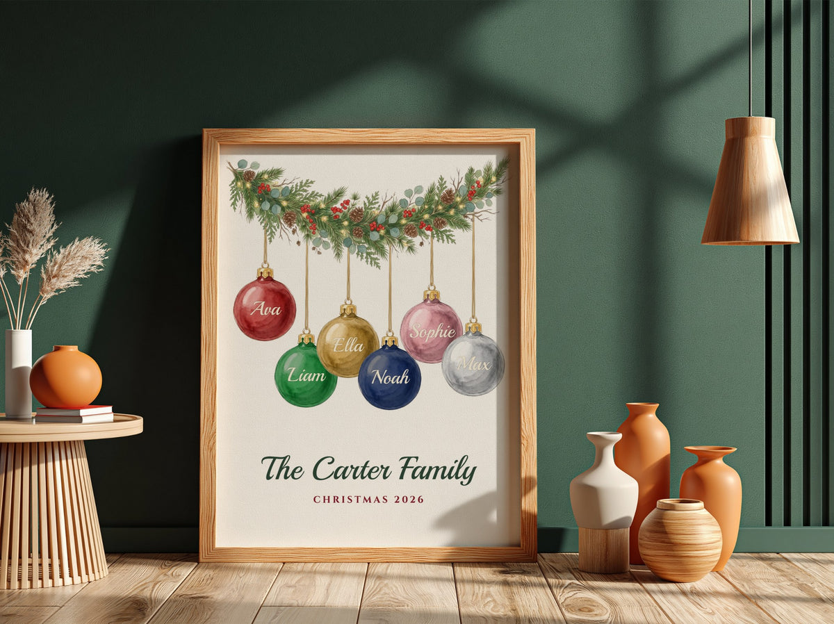 A framed personalized Christmas art print leaning against a dark green wall on a wooden floor, surrounded by decorative vases and dried grass. The print shows colorful hanging ornaments with the names 'Ava', 'Liam', 'Noah', and 'Sage' above the text 'The Carter Family' and 'Christmas 2026'.