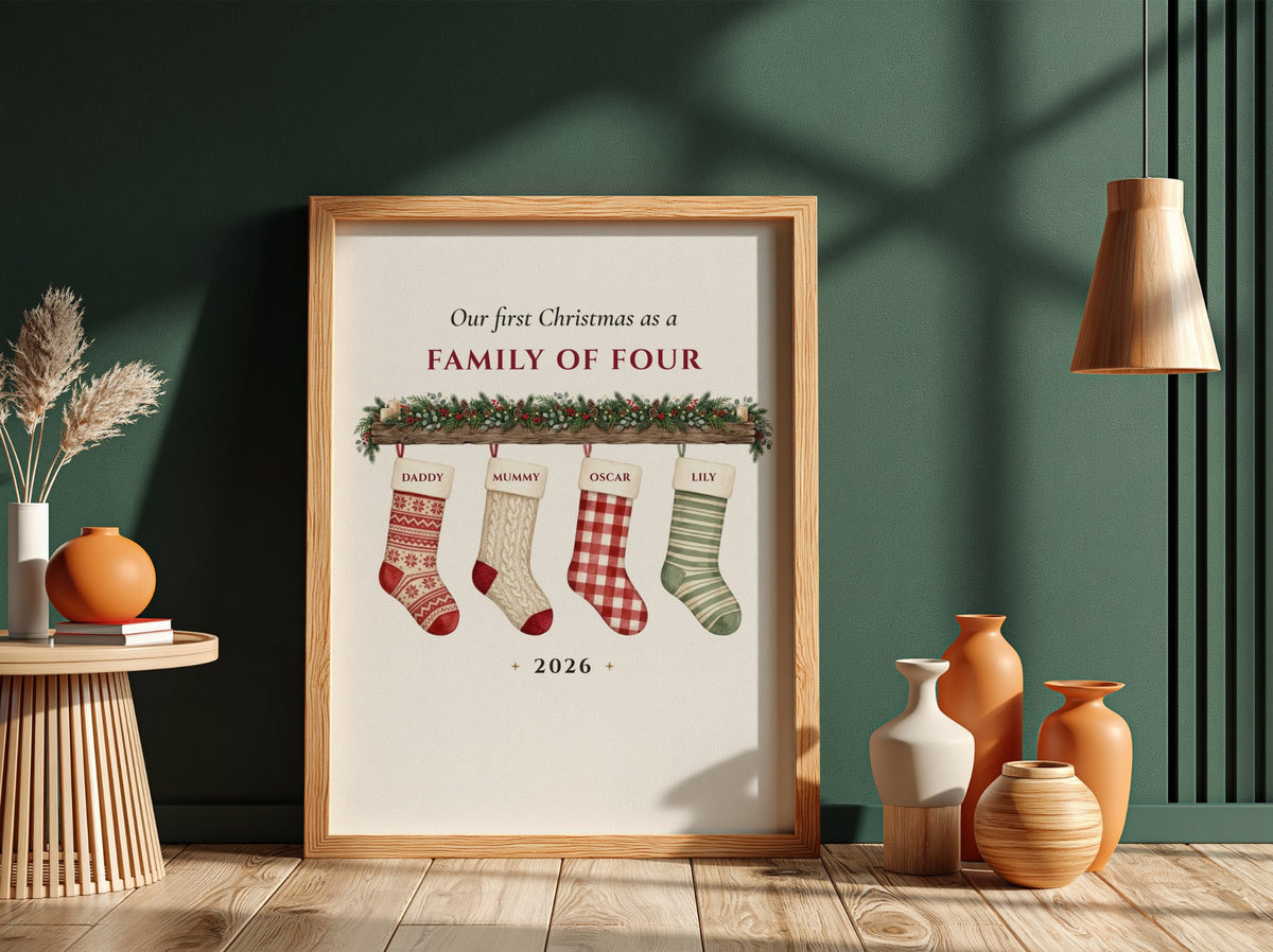 A framed personalized Christmas print leaning against a dark green wall on a wooden floor, surrounded by decorative vases and dried grass. The print reads 'Our first Christmas as a FAMILY OF FOUR' with four hanging stockings and the year 2026.