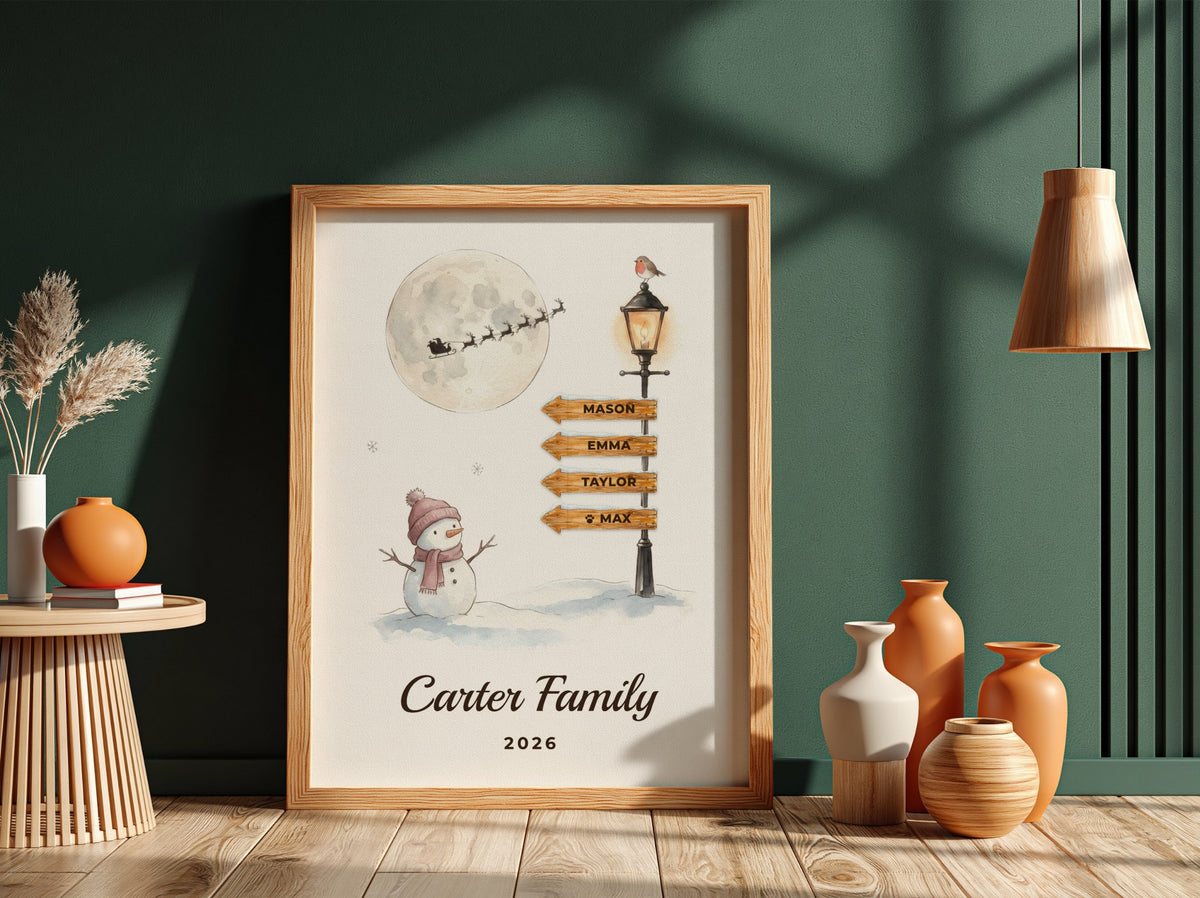 A framed personalized winter-themed print leaning against a dark green wall on a wood floor, surrounded by decorative vases and dried grass. The print shows a snowman, a streetlamp with personalized names on signs, a full moon with Santa's sleigh, and the text 'Carter Family 2026'.